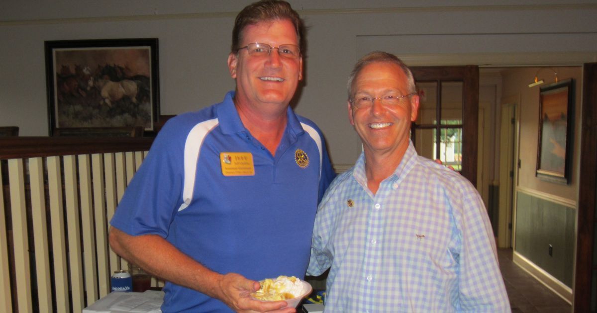 Two smiling men posing indoors, one in a blue polo shirt holding a small dessert.