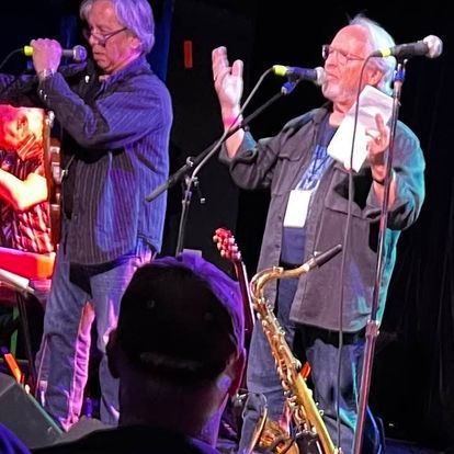 A man singing into a microphone while another man plays a saxophone