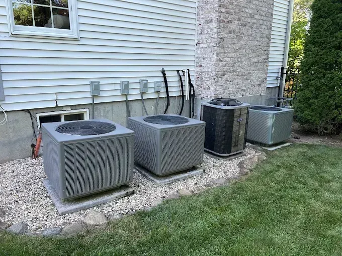 Four outdoor air conditioning units on concrete pads next to a house with a grass lawn.