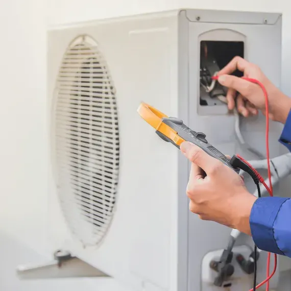Person using a multimeter to test an air conditioner unit's wiring, outdoors.