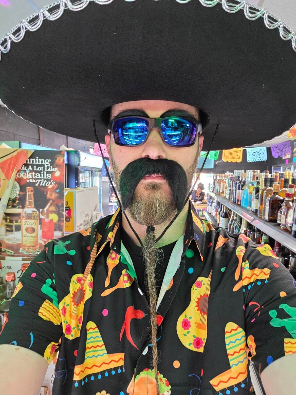 Person in a black sombrero and reflective sunglasses, wearing a colorful patterned shirt, at an indoor market.