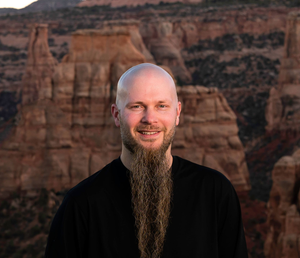 Smiling bald man with a long beard in a black shirt, standing before red canyon cliffs.