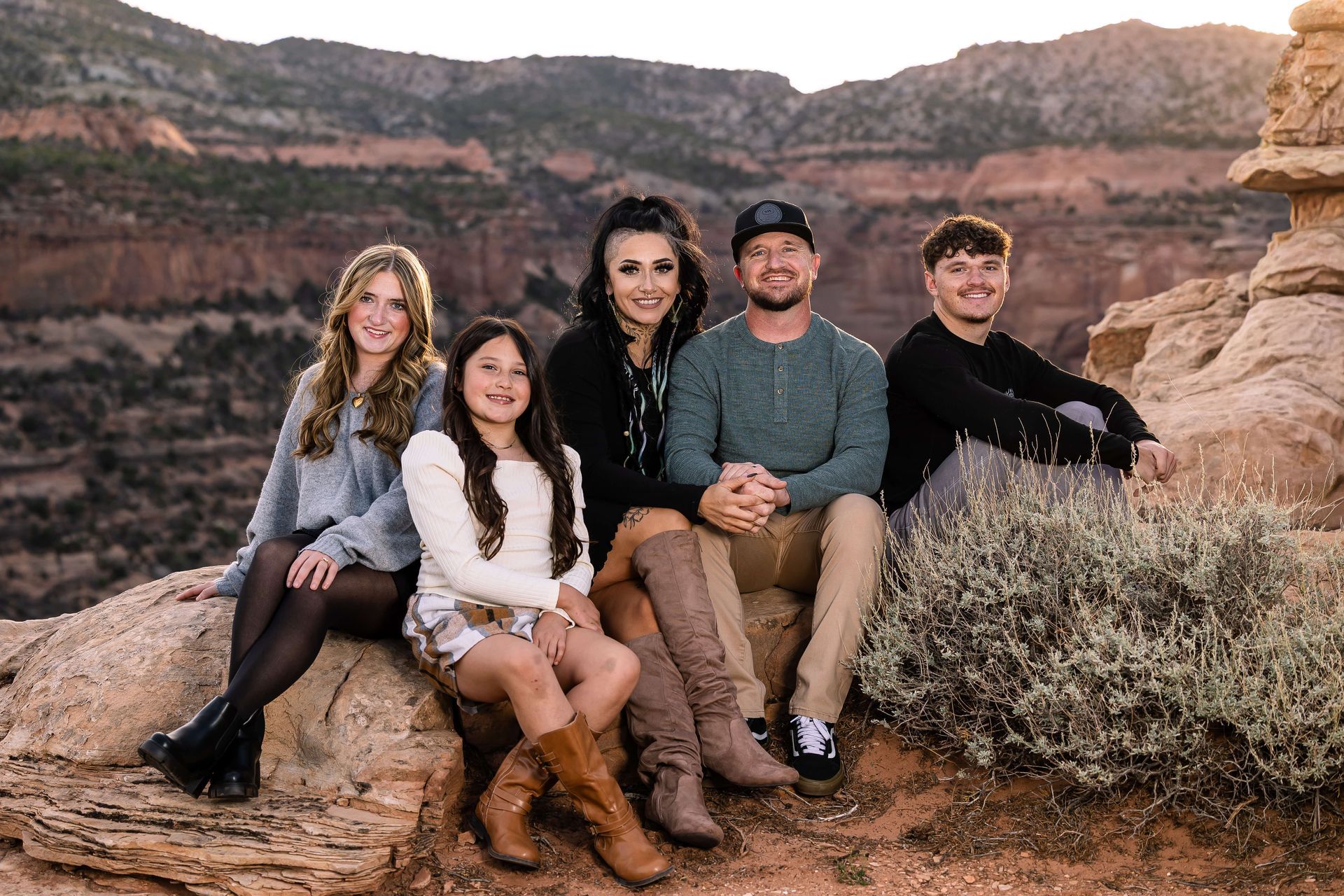 Five people sitting on a rock ledge in a canyon at sunset, smiling at the camera