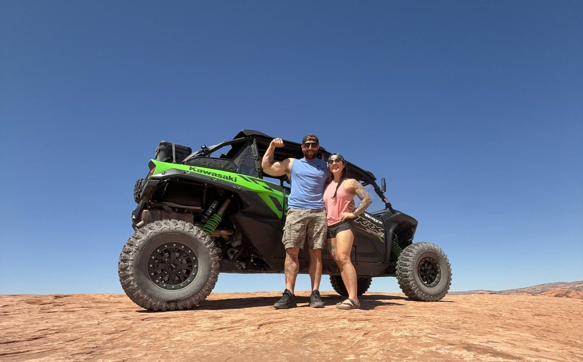 Two people standing by a green-and-black off-road vehicle on a rocky desert hill under a clear blue sky