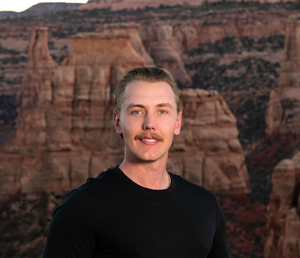 Man in black shirt standing before red canyon cliffs at sunset