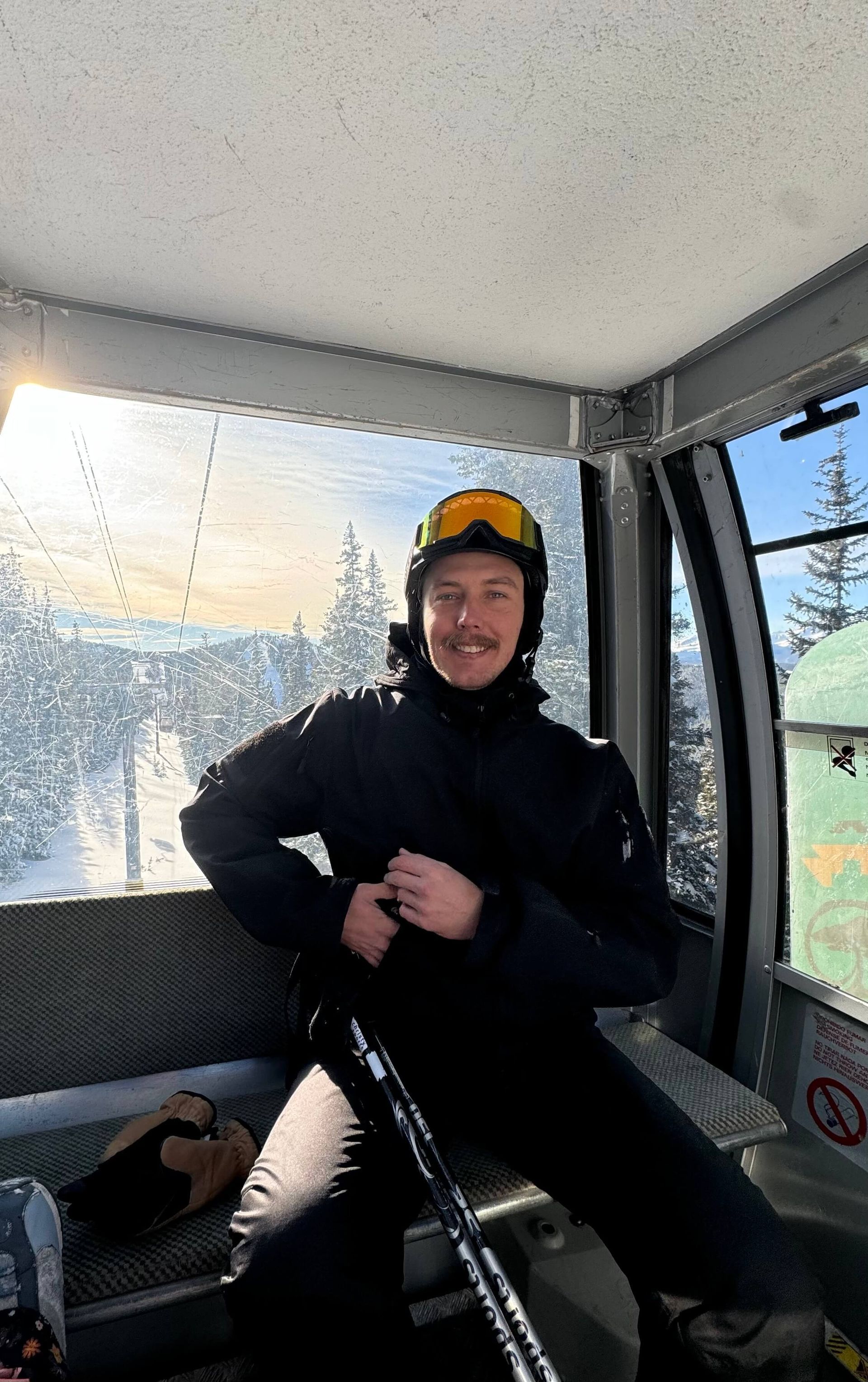 Person in a yellow hard hat and black jacket seated inside a work cab, smiling at the camera.