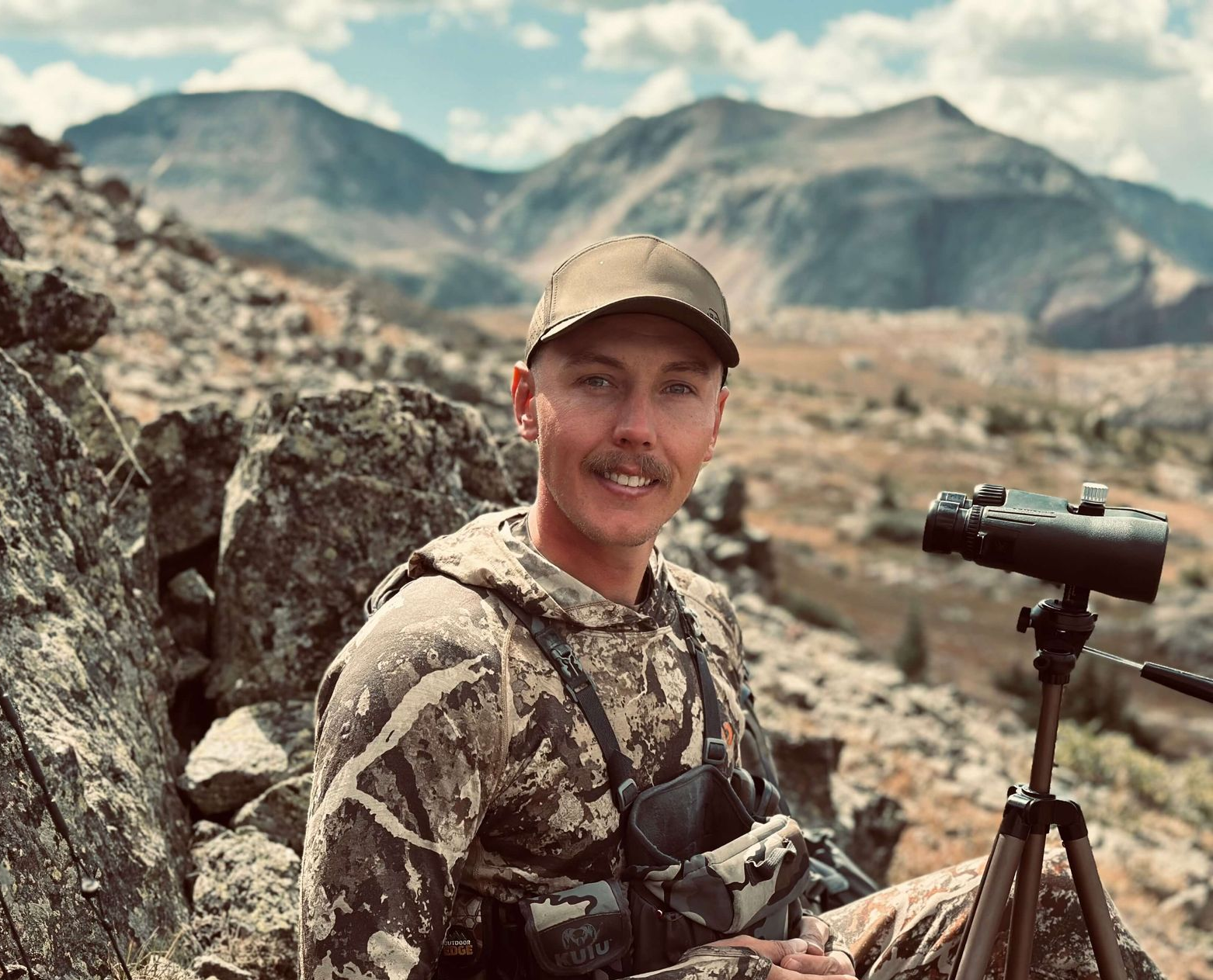 Person in camouflage sits by a tripod-mounted telescope in rocky mountains under a cloudy sky.