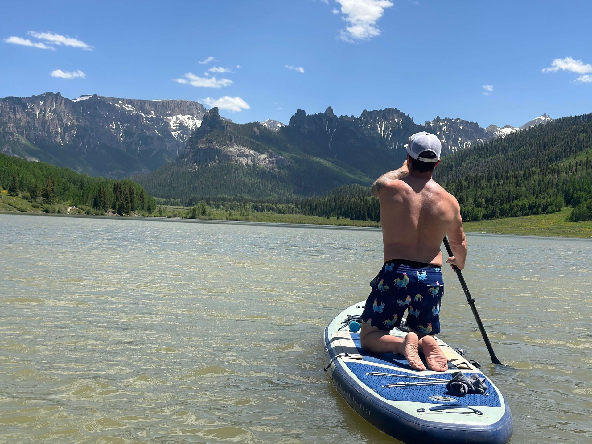 Person paddleboarding on a lake with mountains and trees under a blue sky.