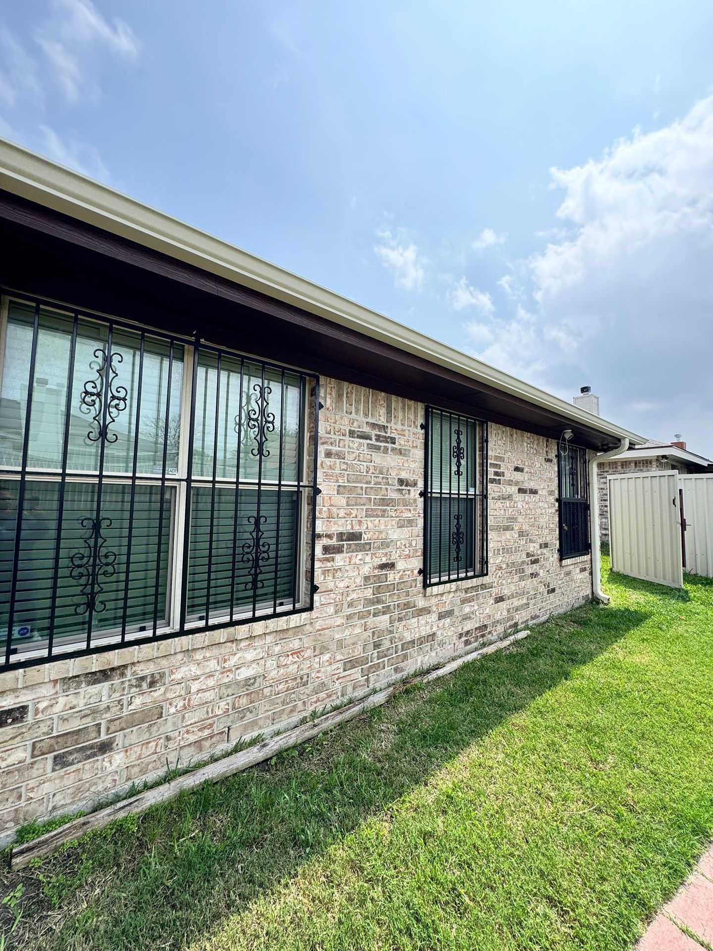 Single-story brick house exterior with barred windows, green lawn, and blue sky