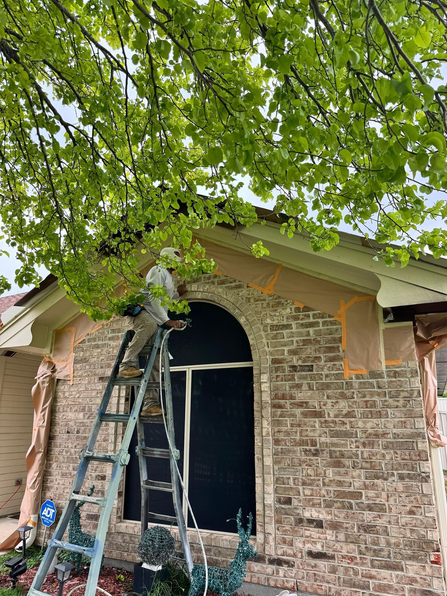 Ladders lean against a brick house under a leafy tree, with a large arched window opening under repair
