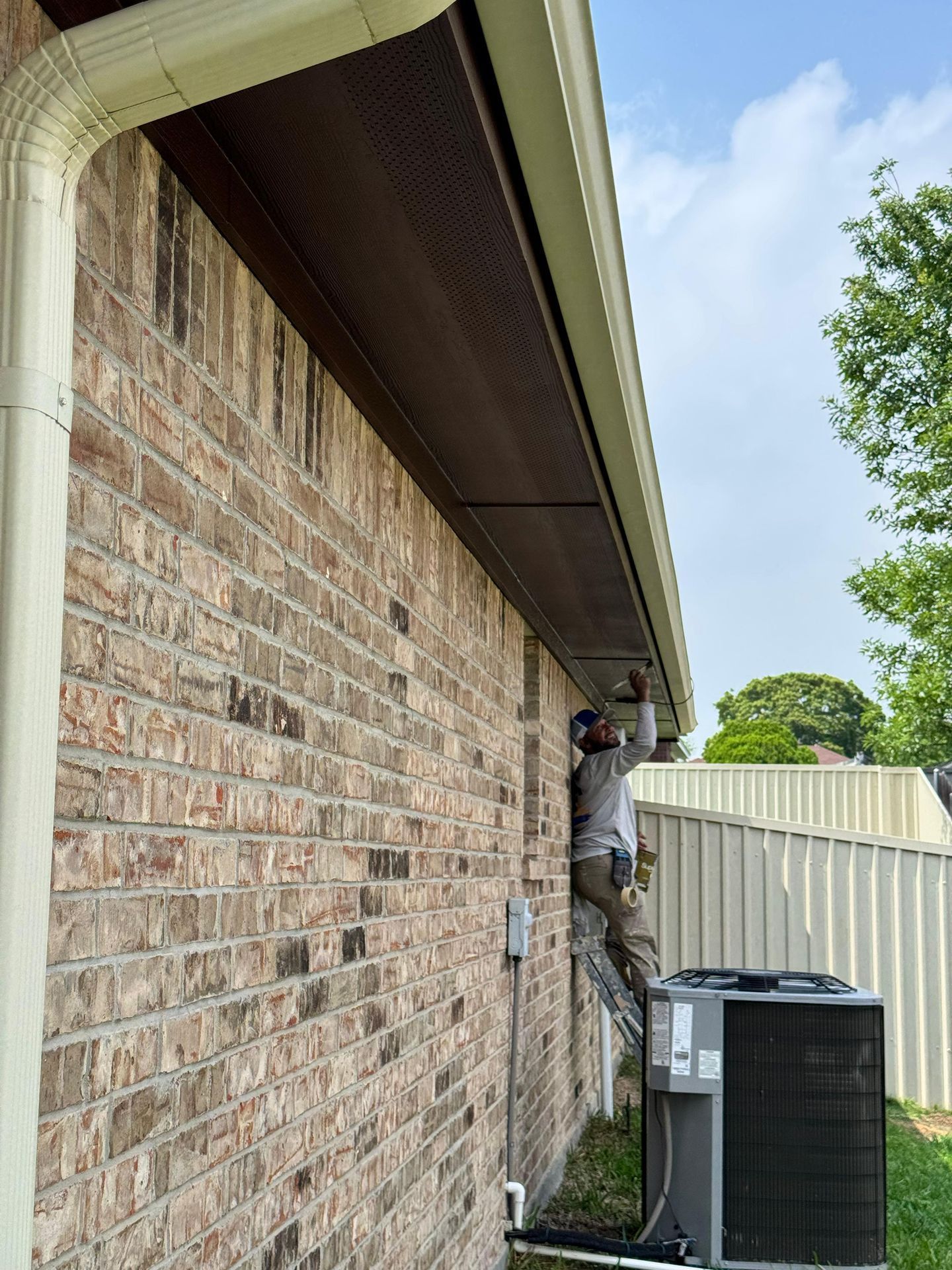 Technician working on an exterior brick wall beside an HVAC unit under a roof eave