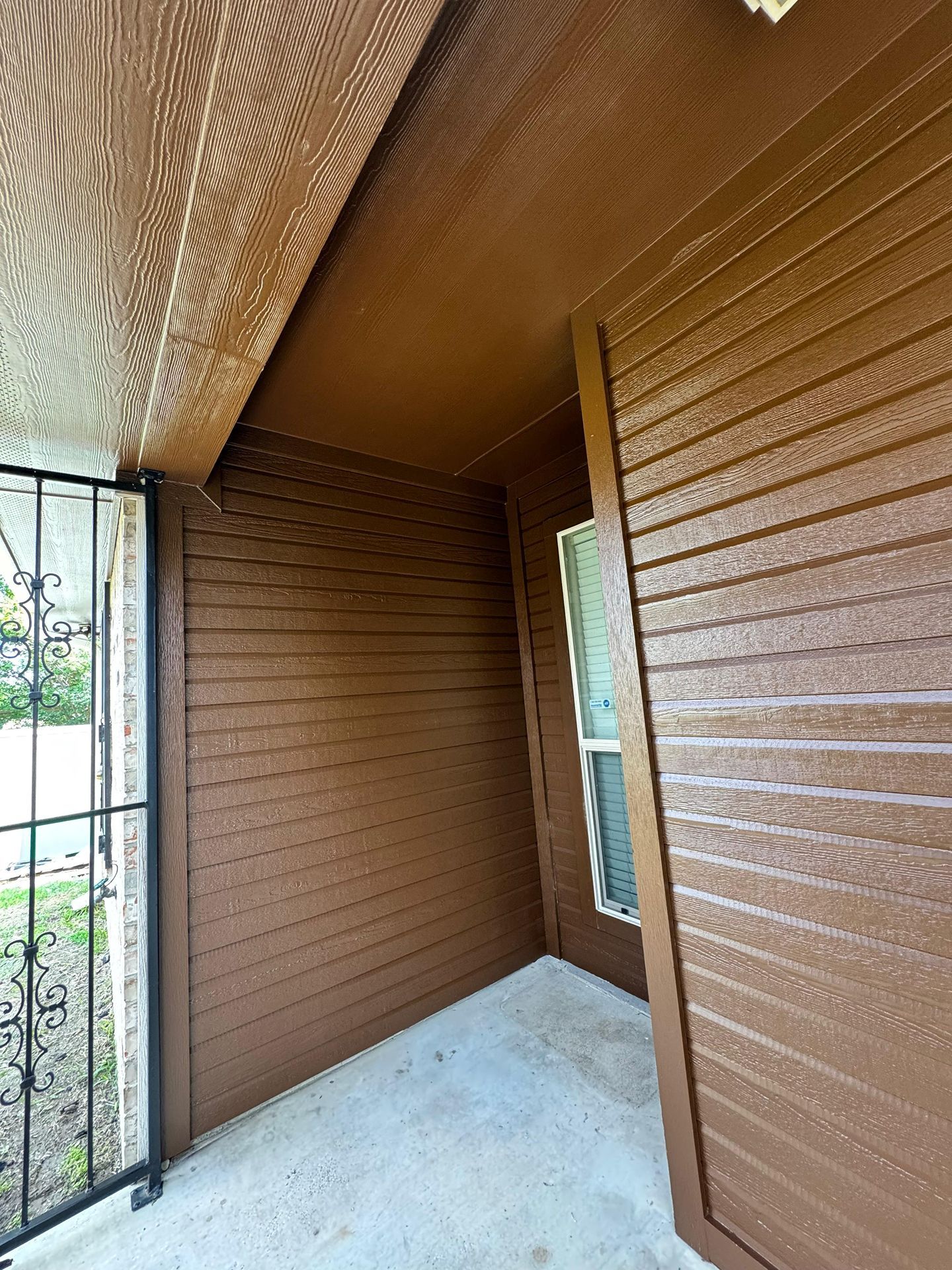 Small covered balcony with brown textured walls, light floor, and a metal railing overlooking greenery.