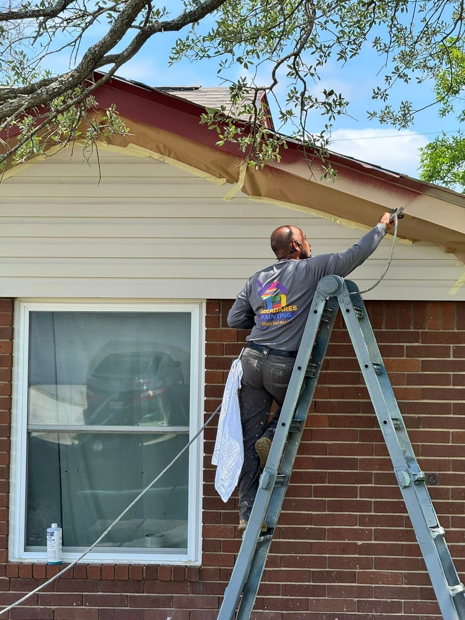 Worker on a ladder painting the trim of a house exterior under a tree.