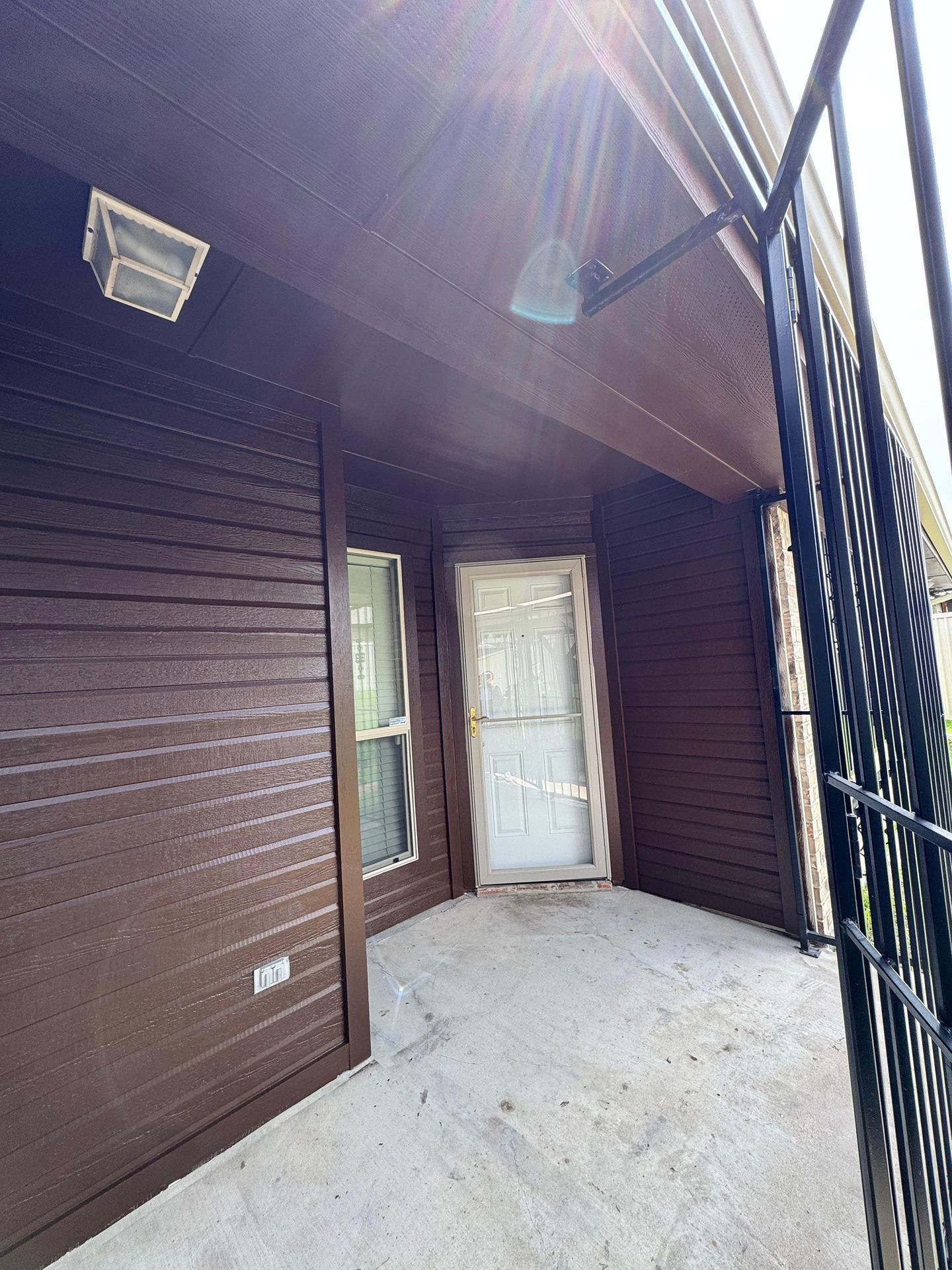 Covered porch with brown wood siding, white door, and black railing in daylight