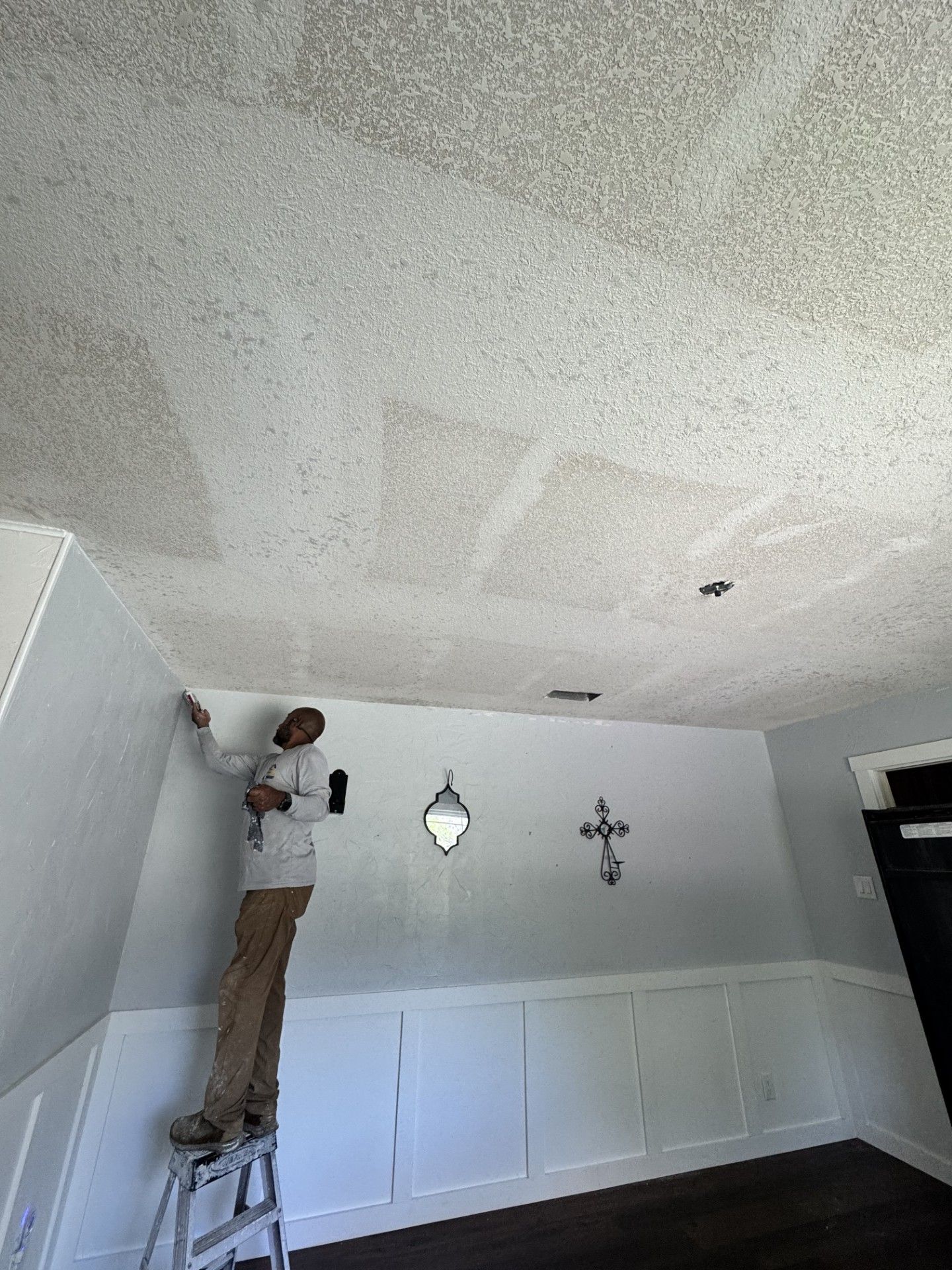 Worker painting a textured ceiling in a dining room with white wainscoting and wall decor.