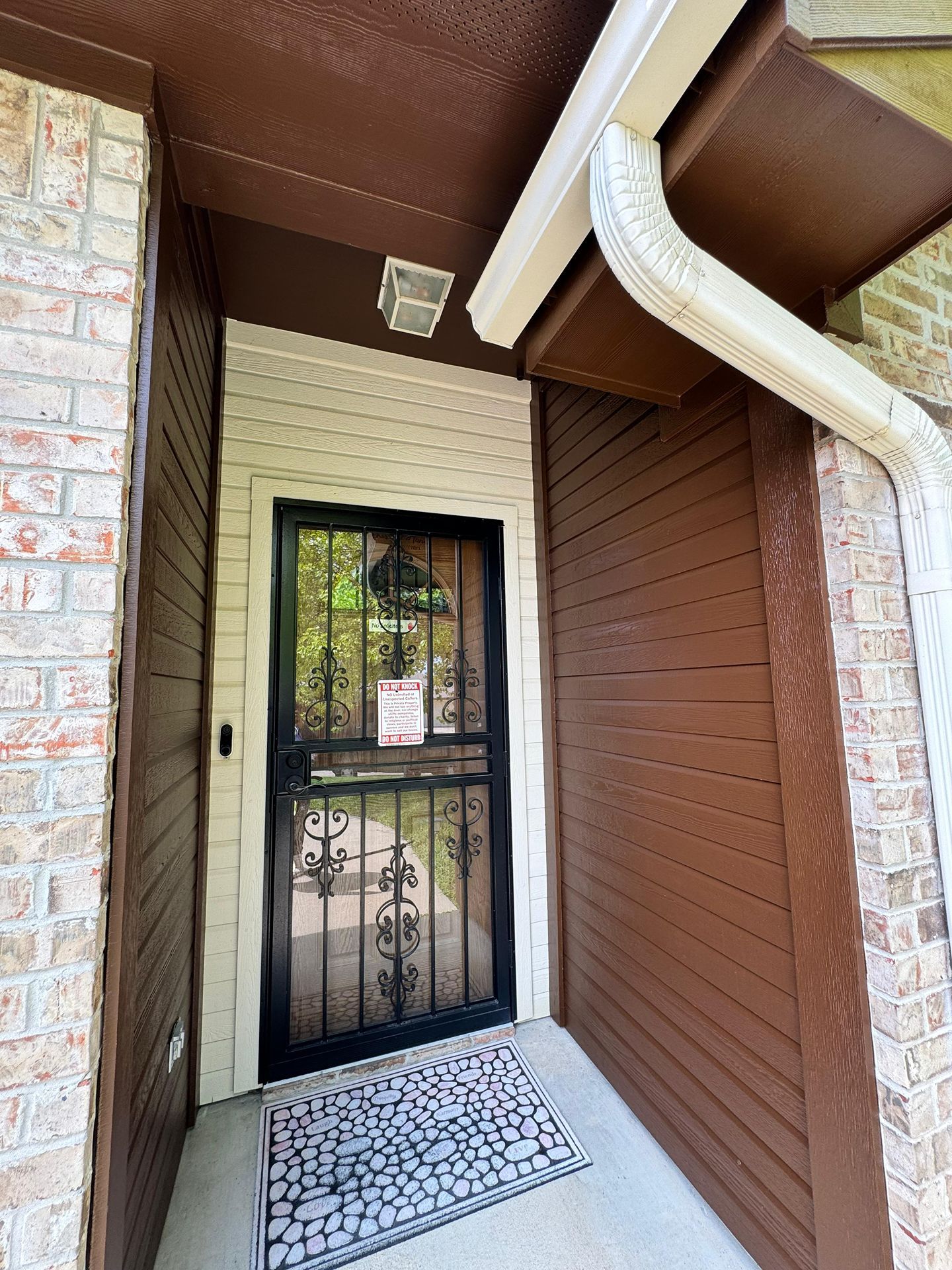 Front entryway with black security door, brick walls, brown trim, and a white downspout under a covered porch