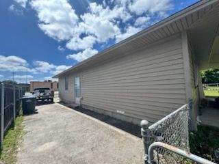 Narrow concrete side yard beside a beige house under a blue sky, with fence, trash bins, and a driveway at the end