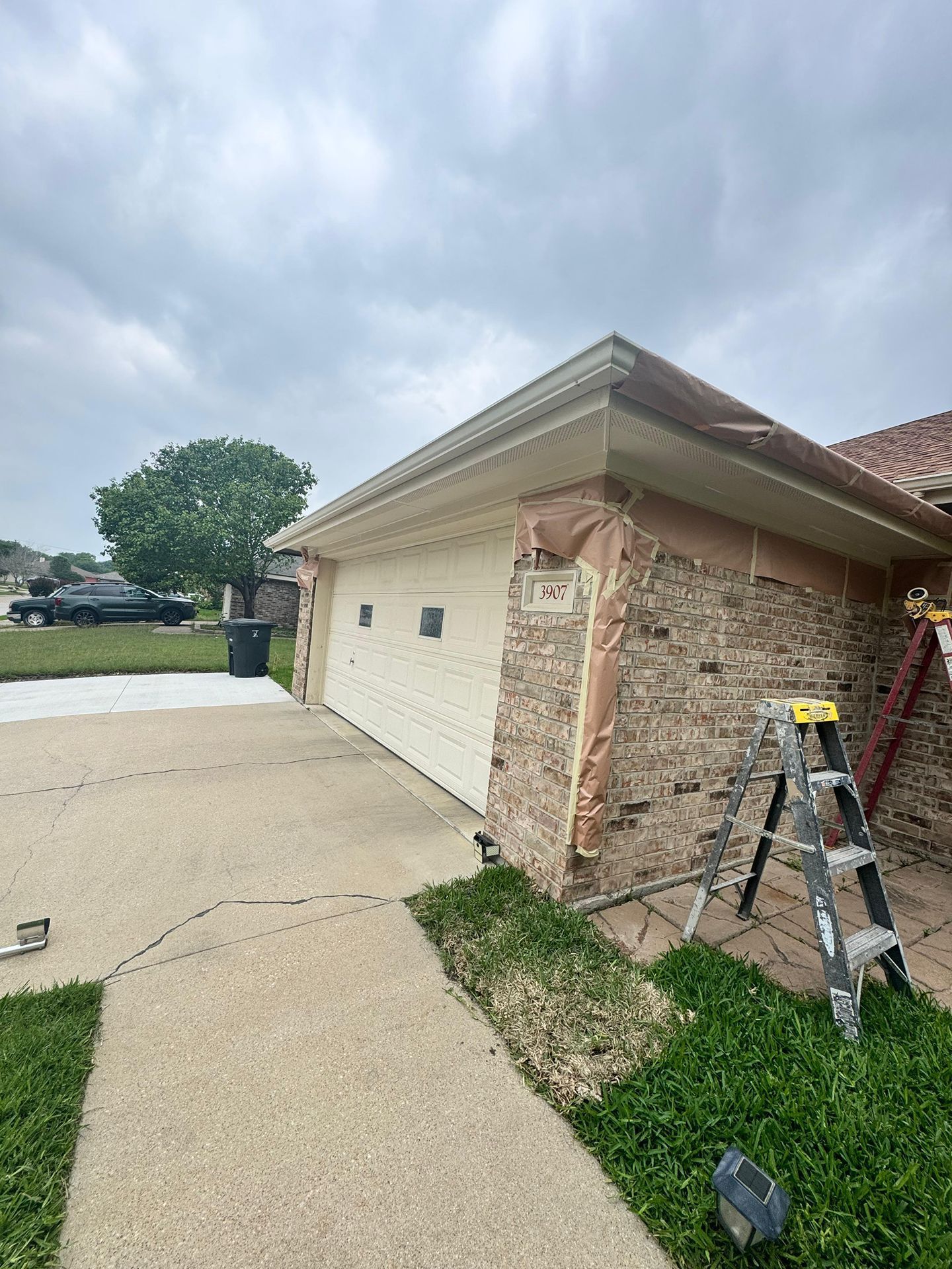 Brick house corner with ladder, concrete driveway, and grassy yard under a cloudy sky
