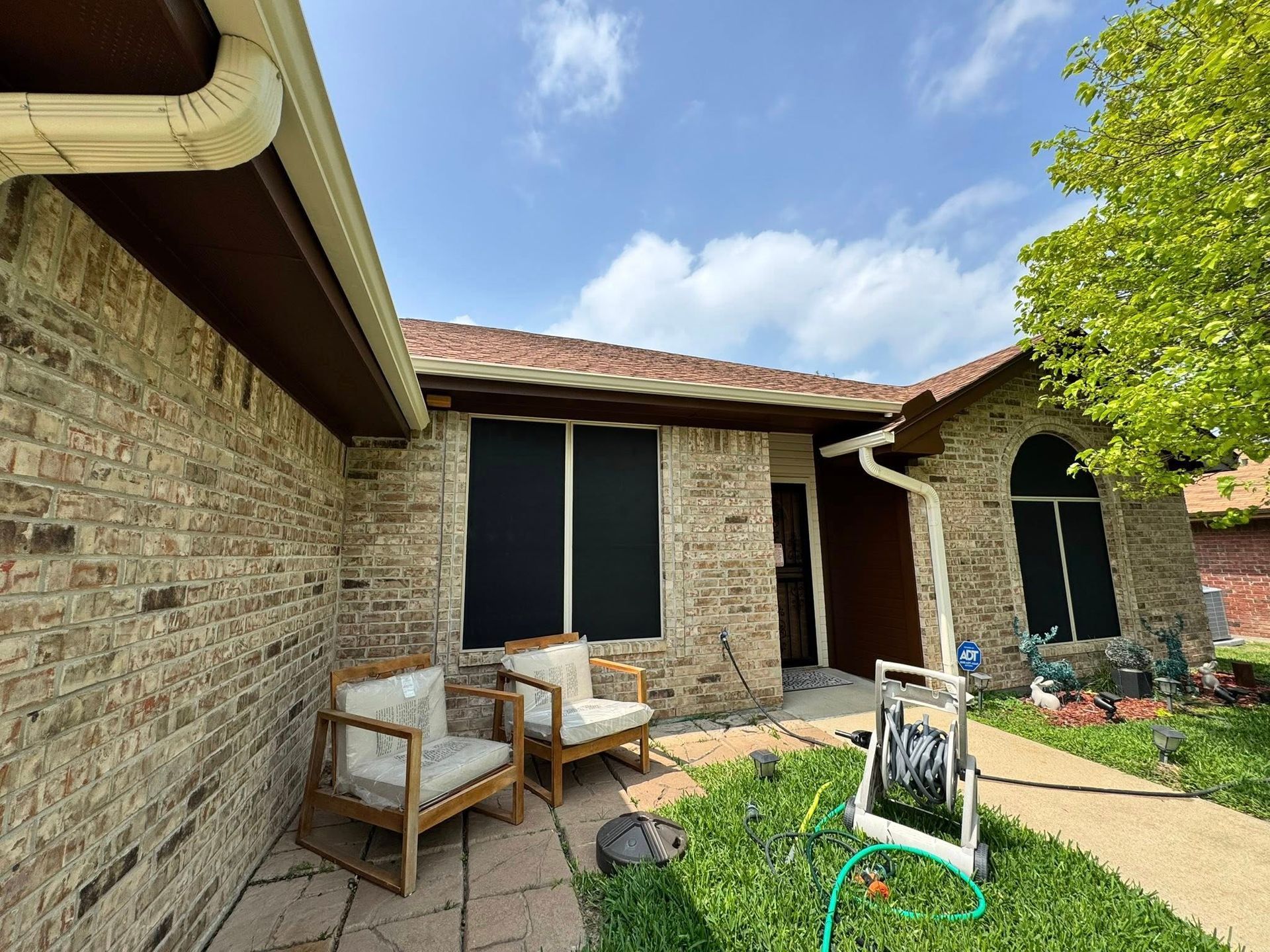 Brick patio with wicker chairs, a small table, and a black sign in front of a house under a blue sky.