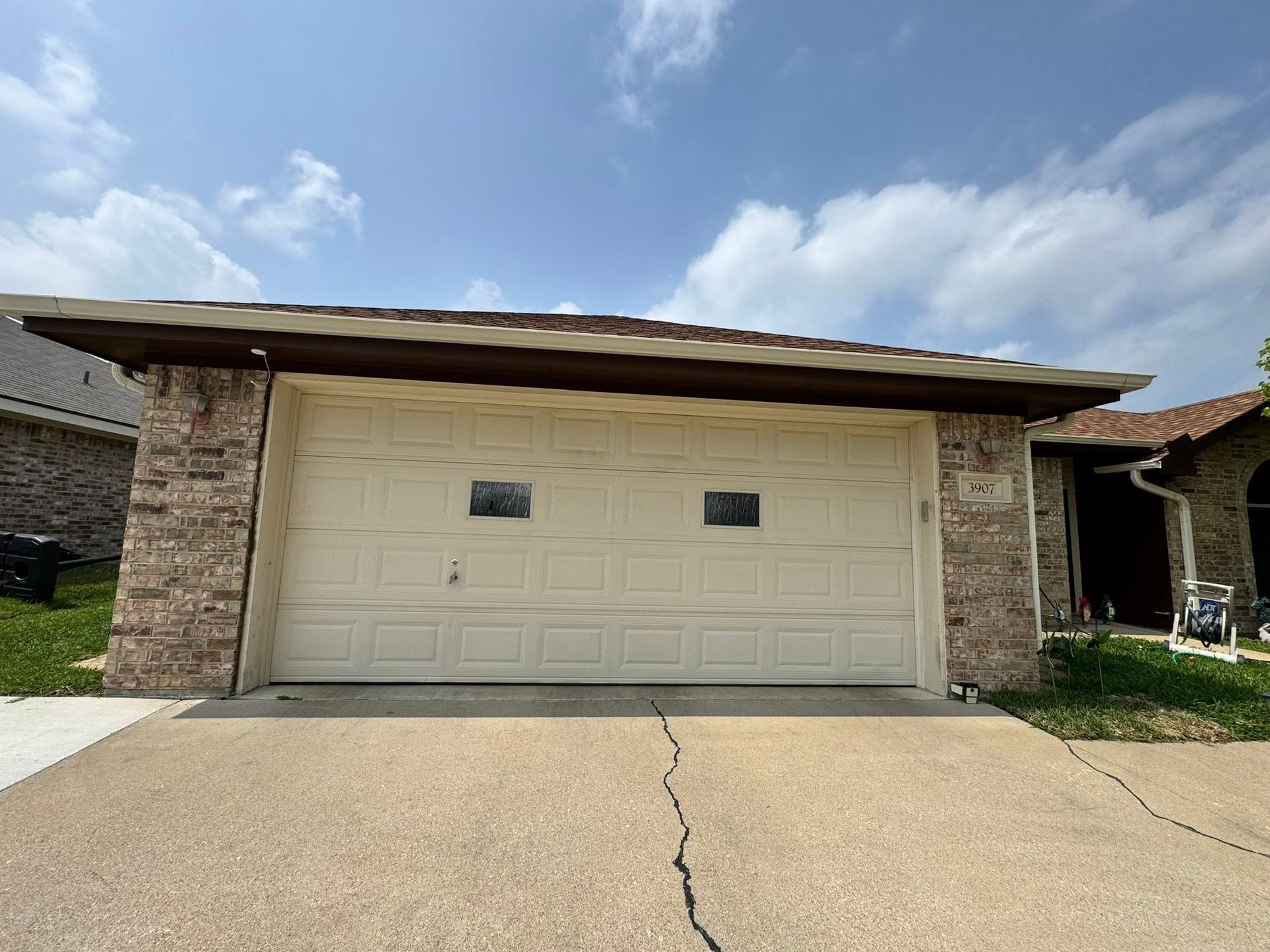 Single-story brick house with a beige double garage door and cracked concrete driveway under a blue sky.
