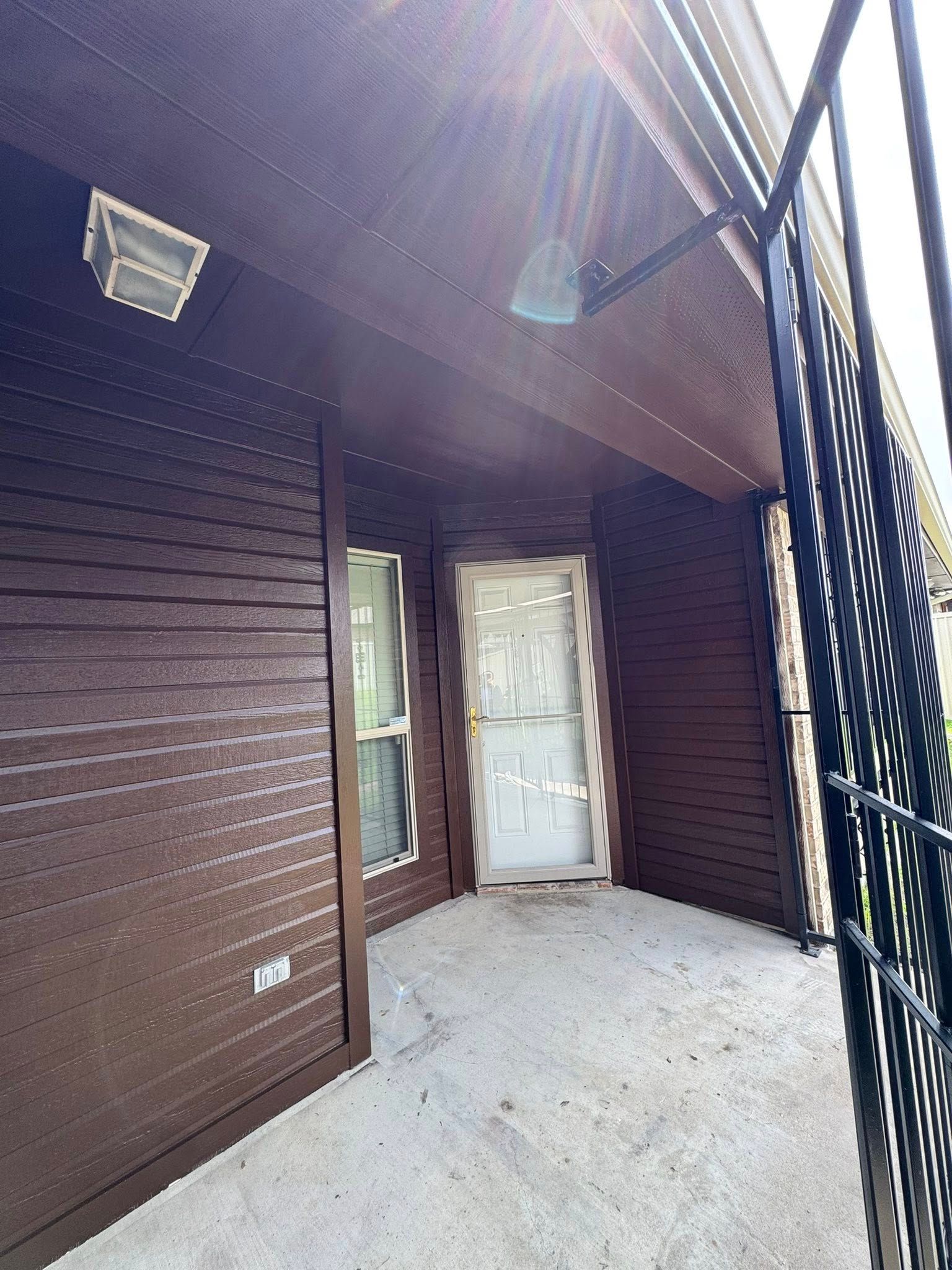 Covered porch with dark wood siding, white door, and black metal railing in bright sunlight