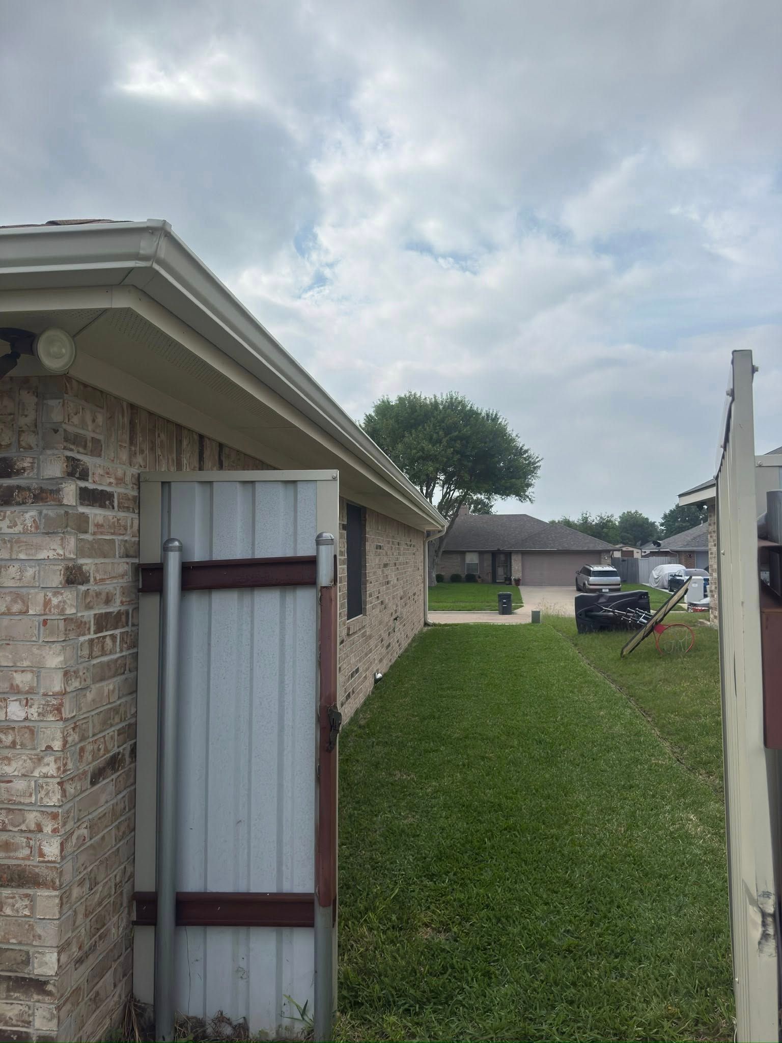 Open side gate between a brick house and a grassy slope, leading to a driveway with a parked car.