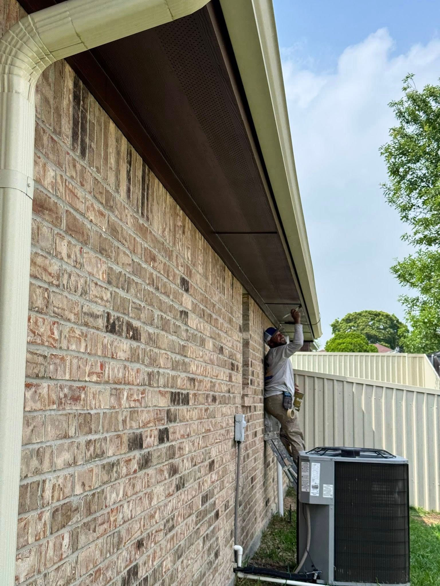 Worker on a ladder repairing exterior brick wall under a roof eave beside an HVAC unit.