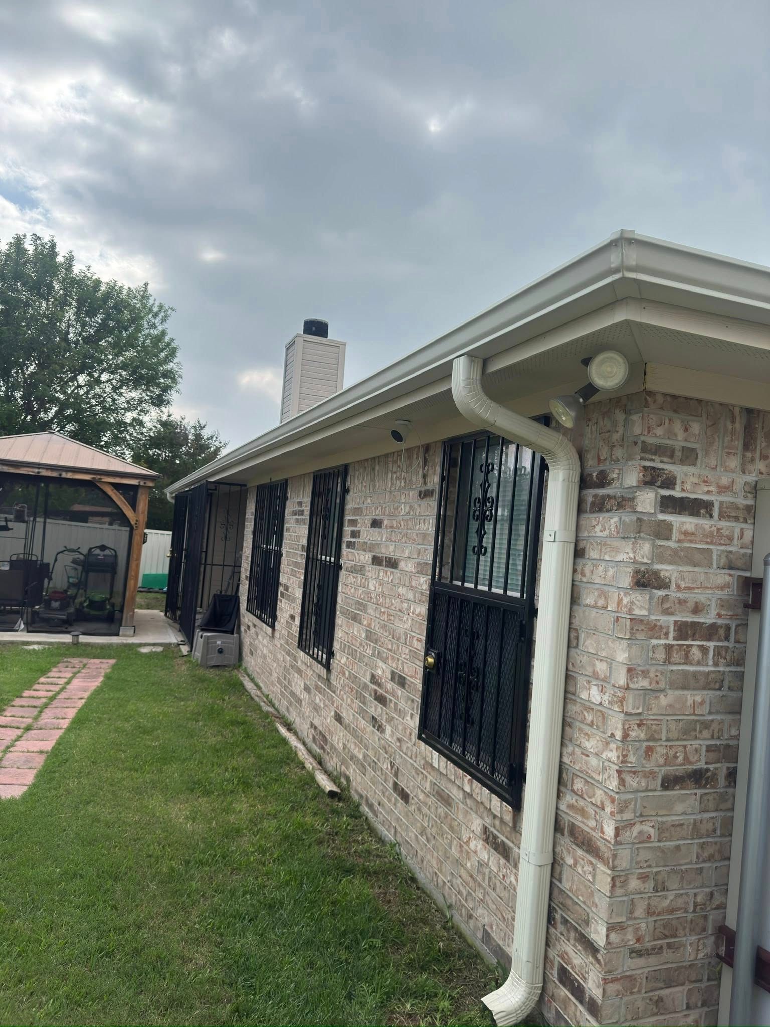 Single-story stone house exterior with black barred windows, white gutter, and grassy yard under cloudy sky