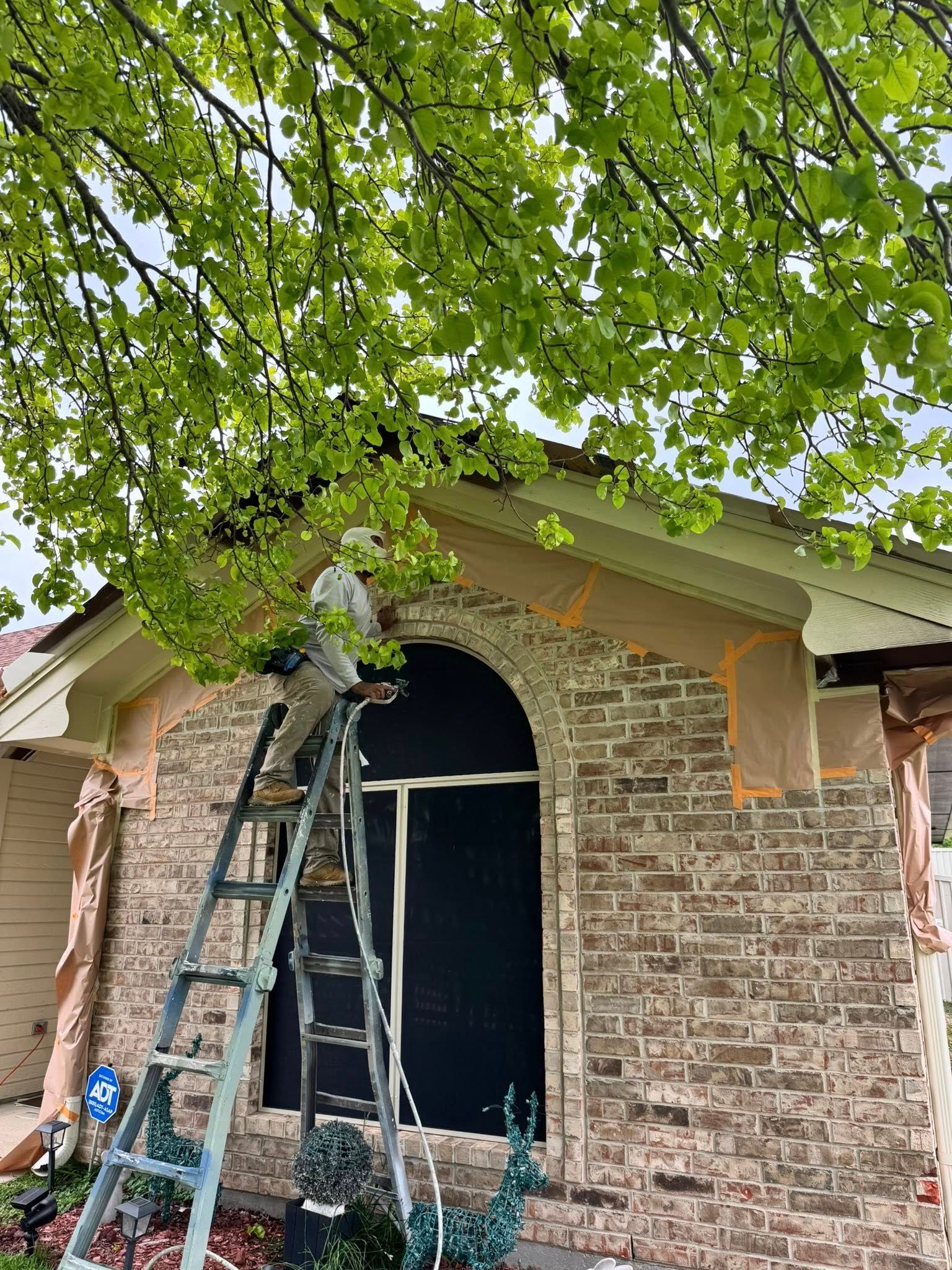 Worker on ladder trimming tree branches above a brick house’s arched front entry.