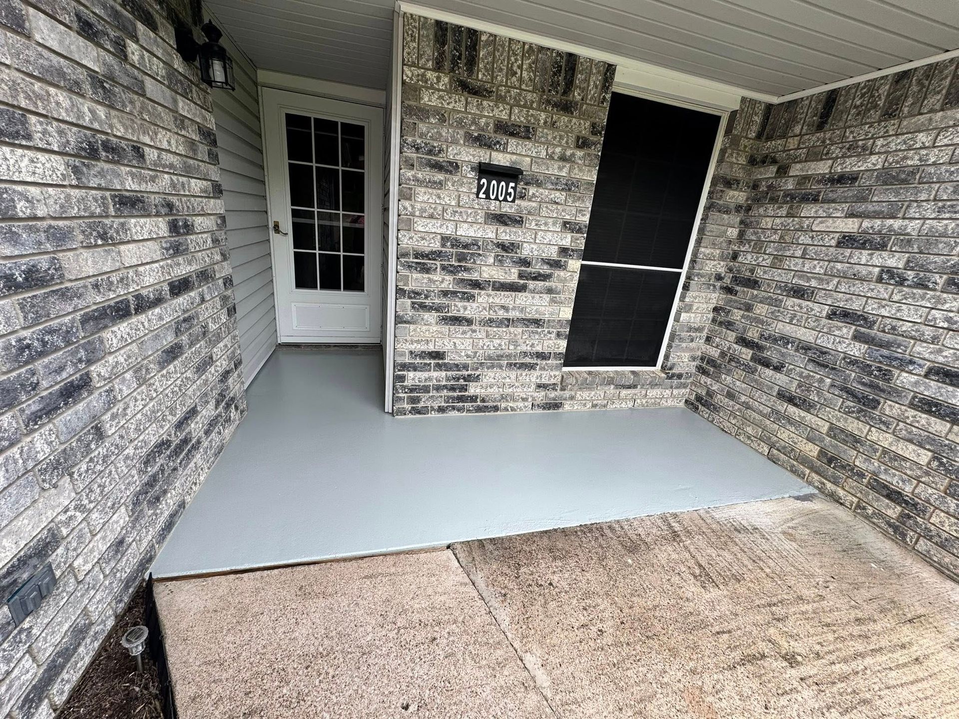 Covered front porch with gray concrete floor, white door, and brick walls.