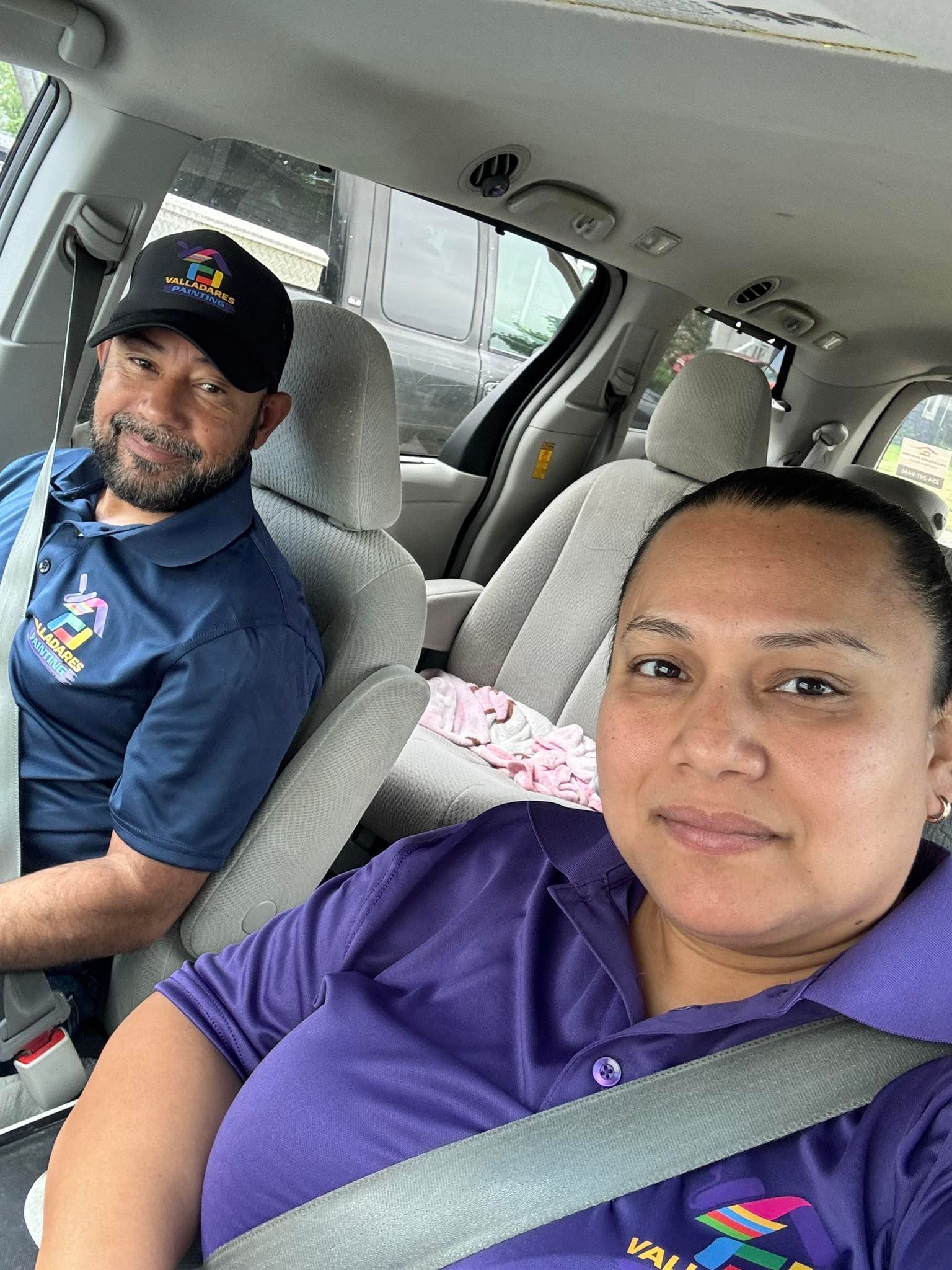Two people seated in a car, wearing purple shirts and seat belts, looking at the camera.