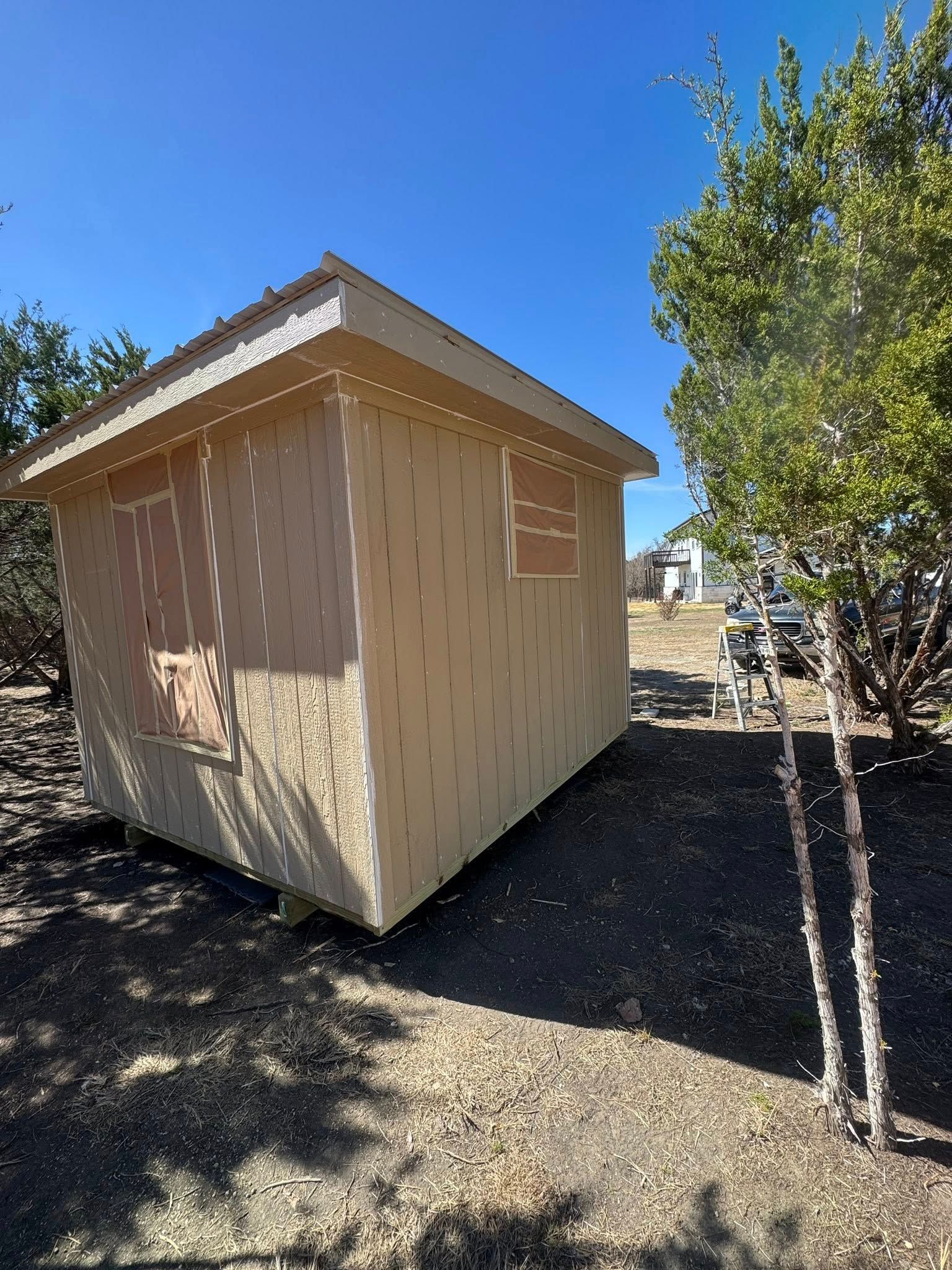 Small tan shed with a pitched roof beside a tree on a sunny day.