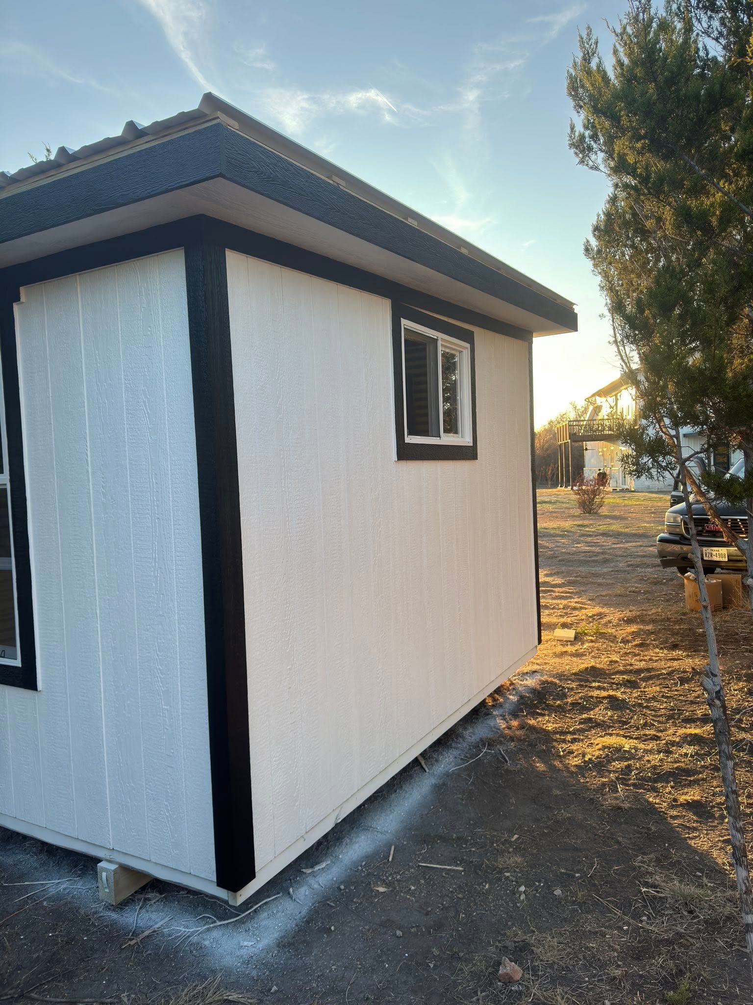 Small white shed with black trim and a window, viewed from the side in a sunny outdoor yard.