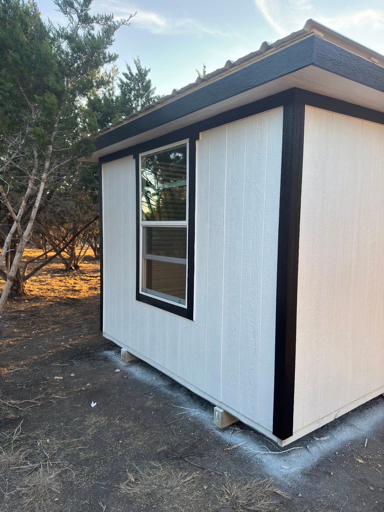 Small white exterior building with black trim and a window, viewed from the side among trees.