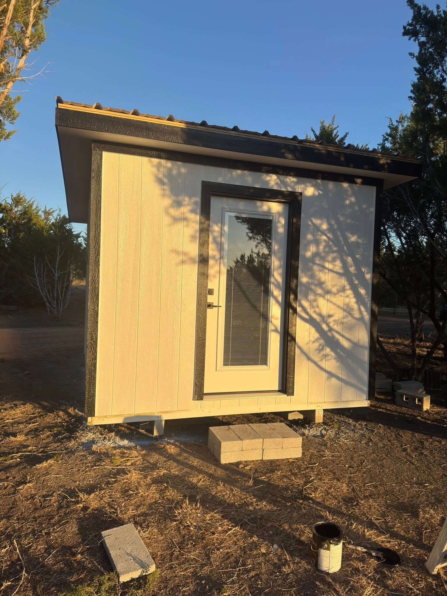 Small beige shed with a slanted roof and white door panel in a gravel yard at sunset