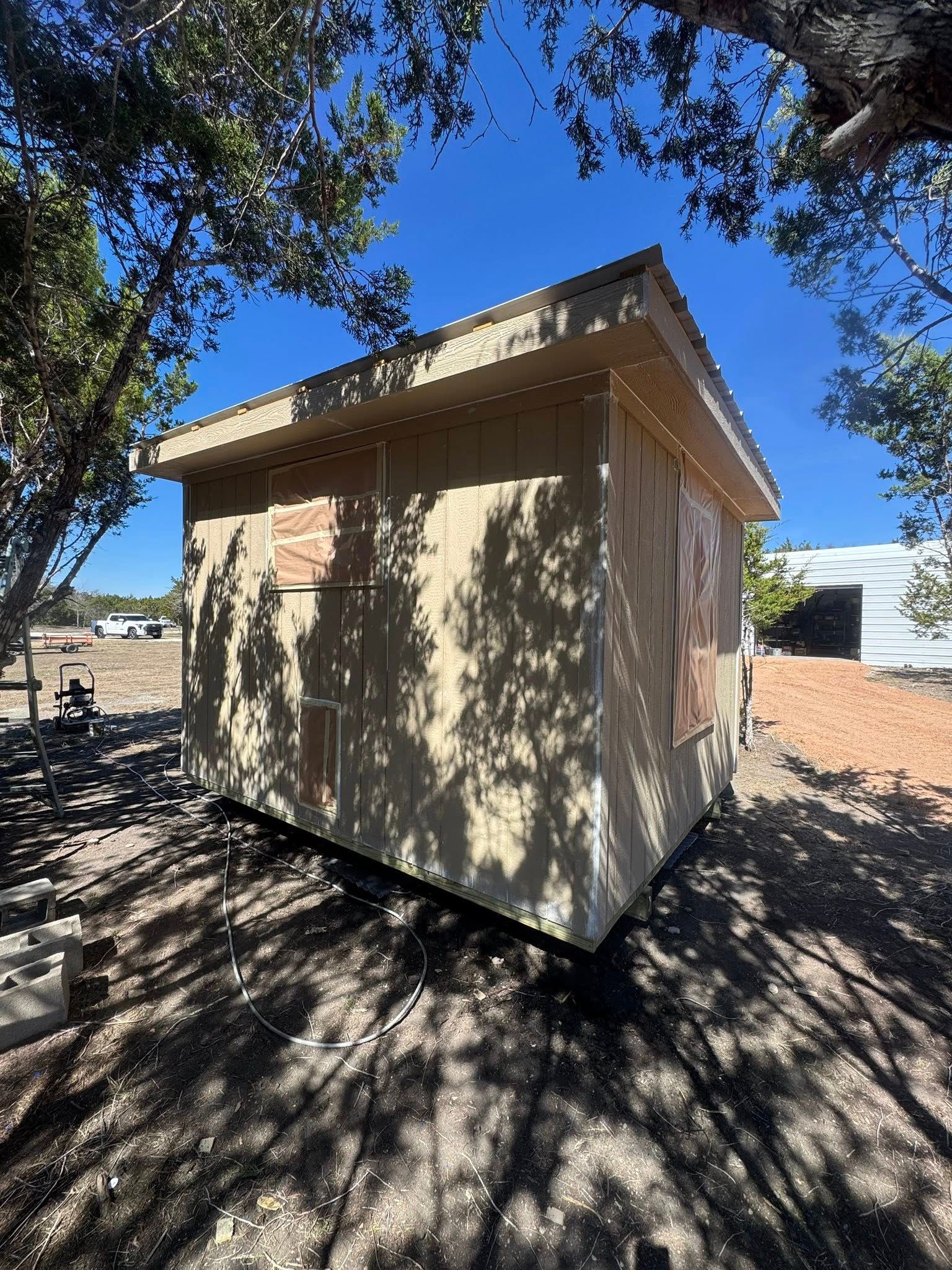 Small tan shed with a slanted roof under trees on a sunny day
