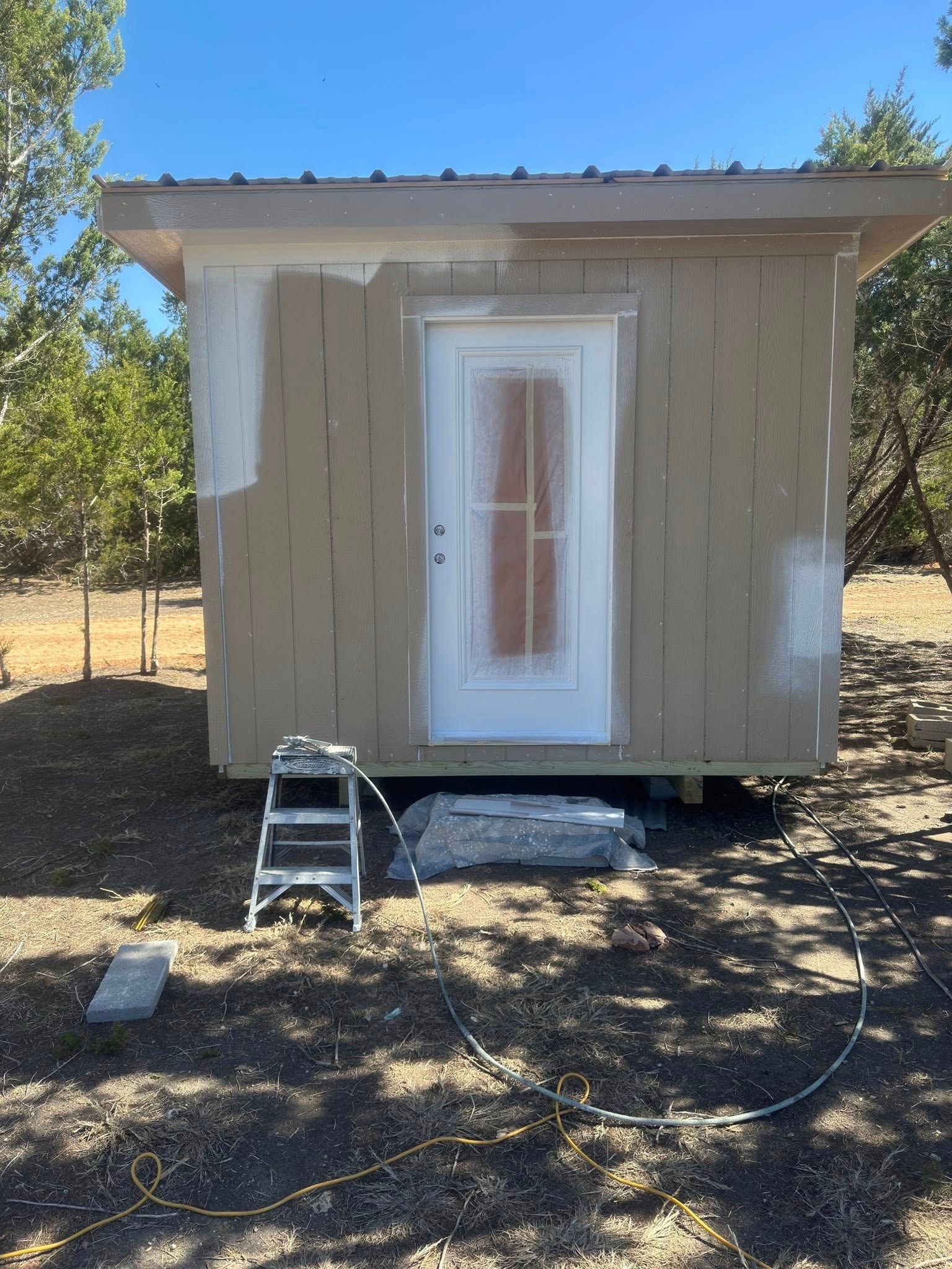 Small tan shed with a white door and a step ladder in front, on a dirt lot under trees.