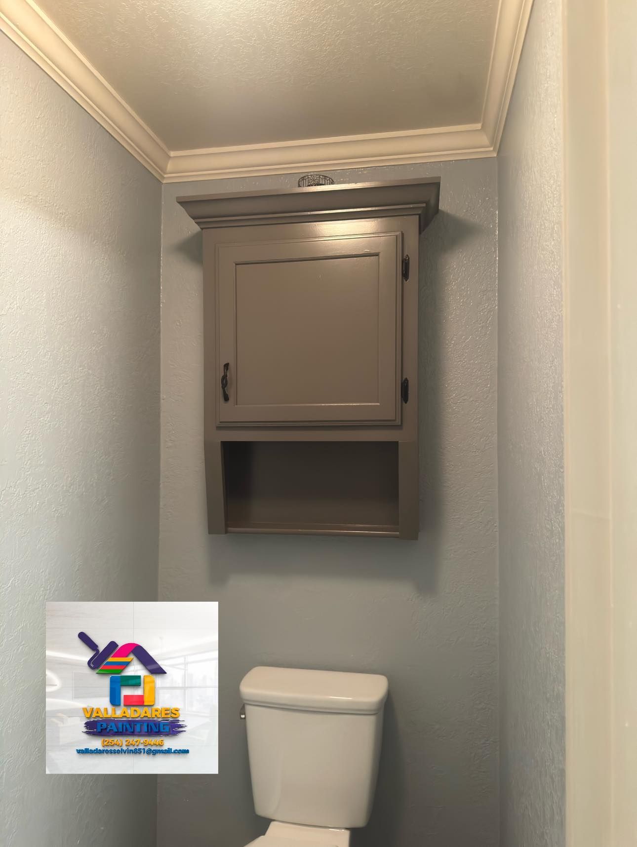 Small bathroom with a wall-mounted cabinet above a toilet and a colorful logo in the lower left.