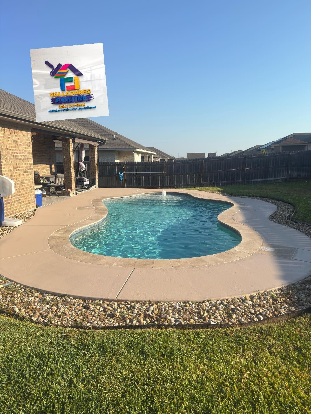 Backyard pool with curving blue water, tan deck, green lawn, and a house under a clear sky