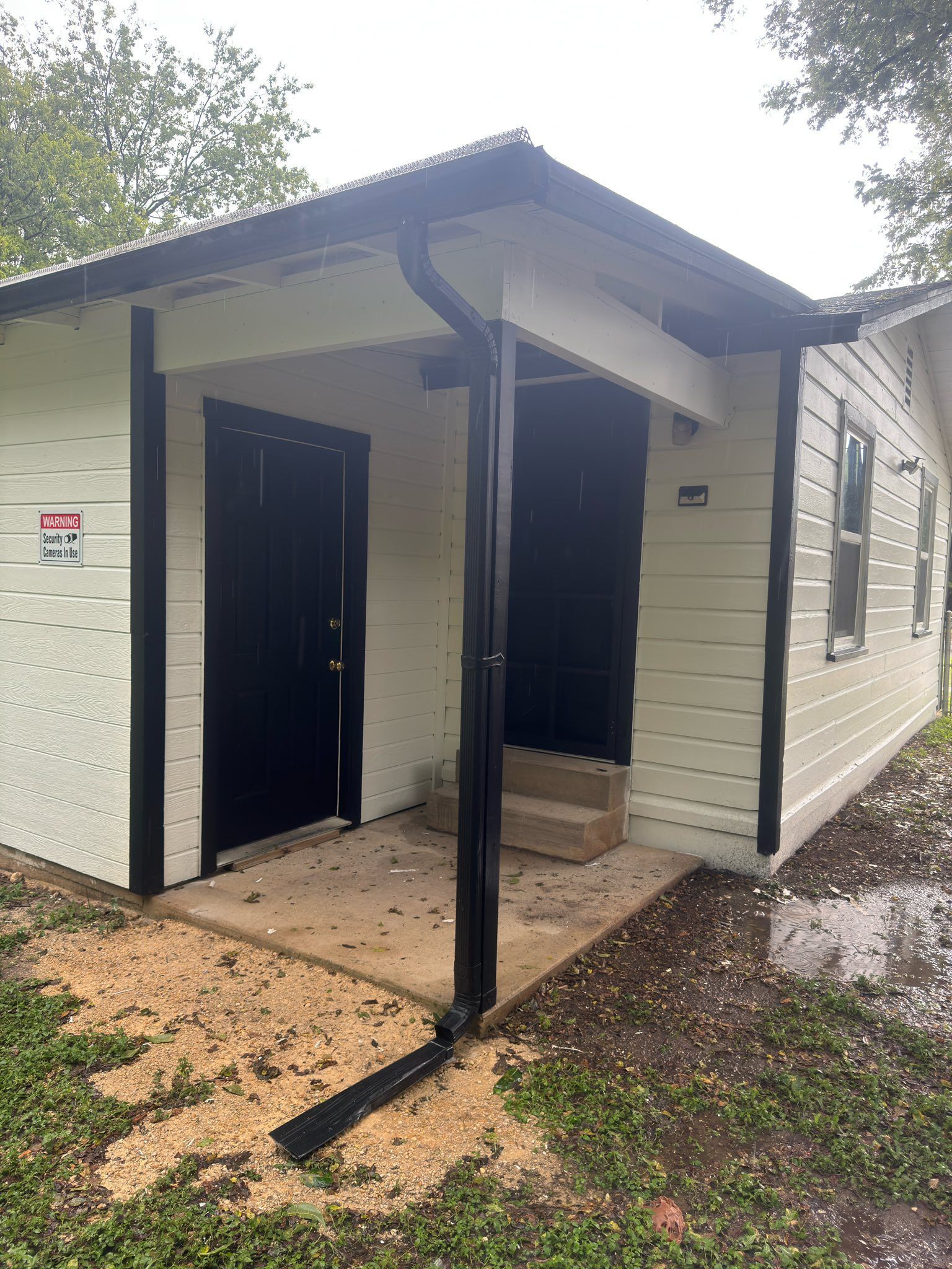 Small white building with black trim, two black doors, and a covered entryway on a dirt path.
