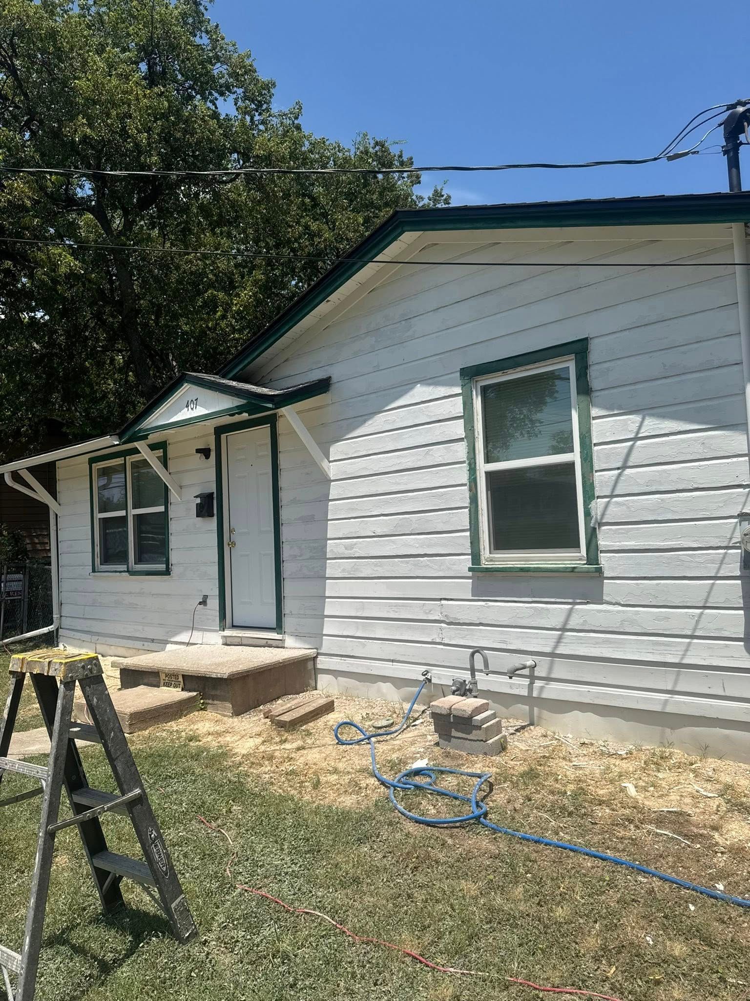 Side of a white house with green trim, a small porch, ladder, and garden hose in a sunny yard.