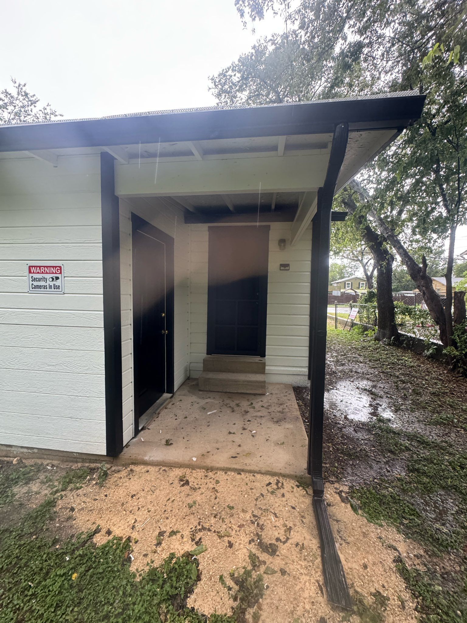 Open doorway of a small white building beside muddy ground and trees after rain.
