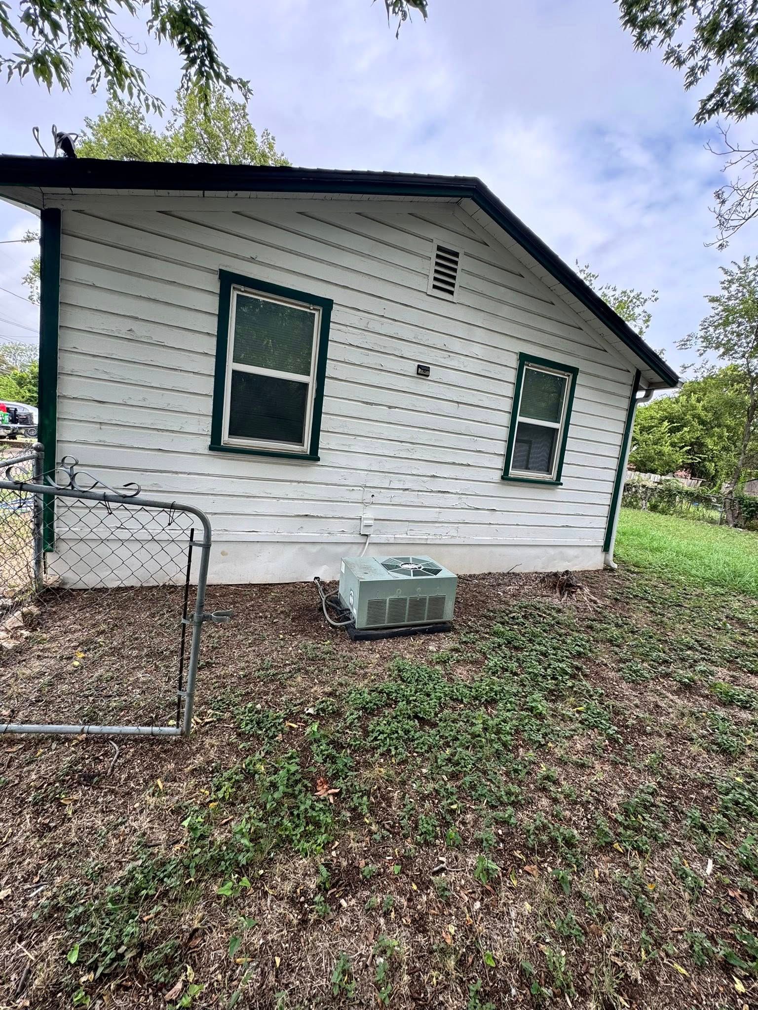 White-sided house with green-trimmed windows, a metal gate, and an outdoor AC unit beside a leaf-covered yard.