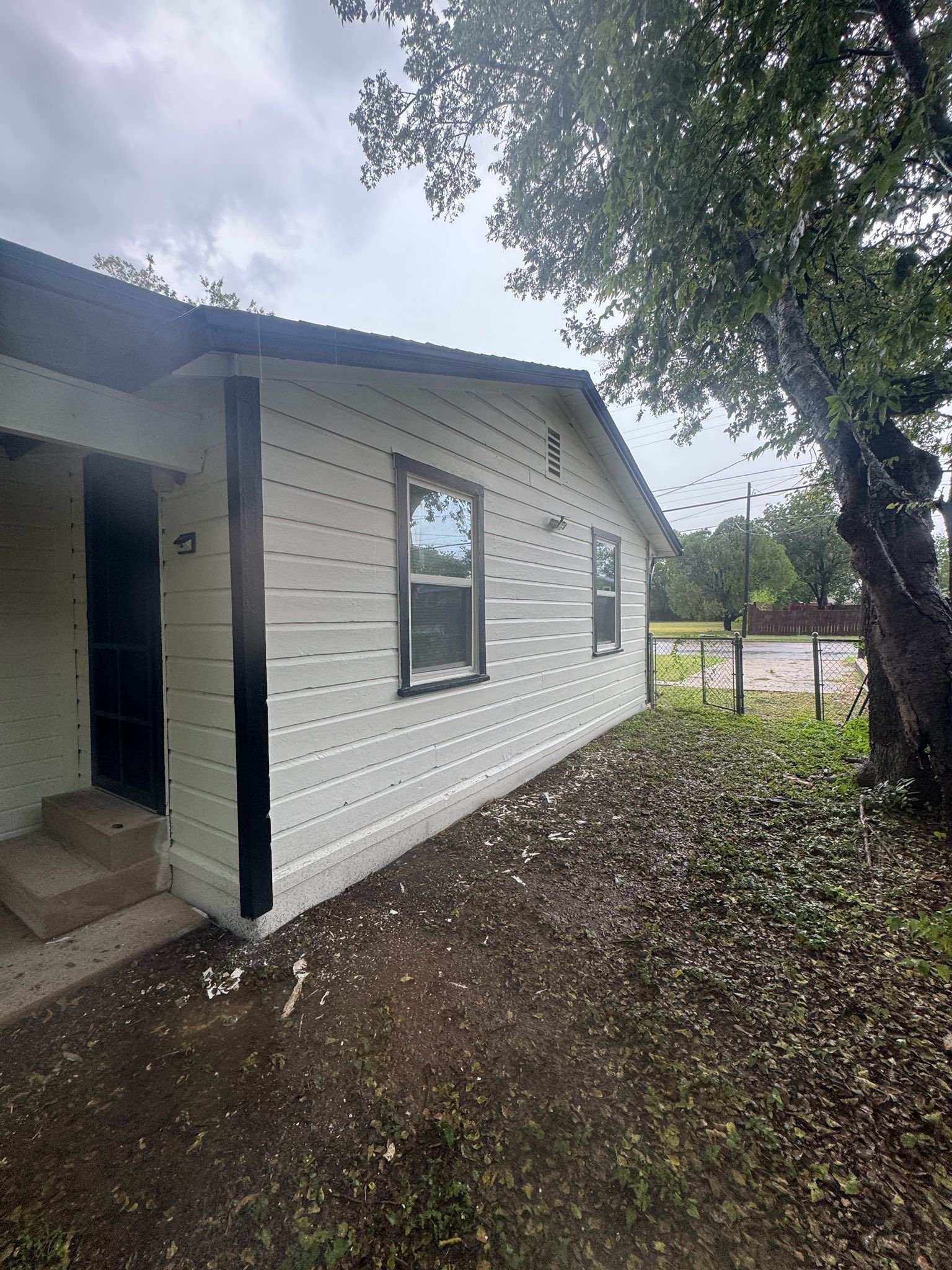 Small white house exterior with a window, porch, and tree overhanging a dirt yard.