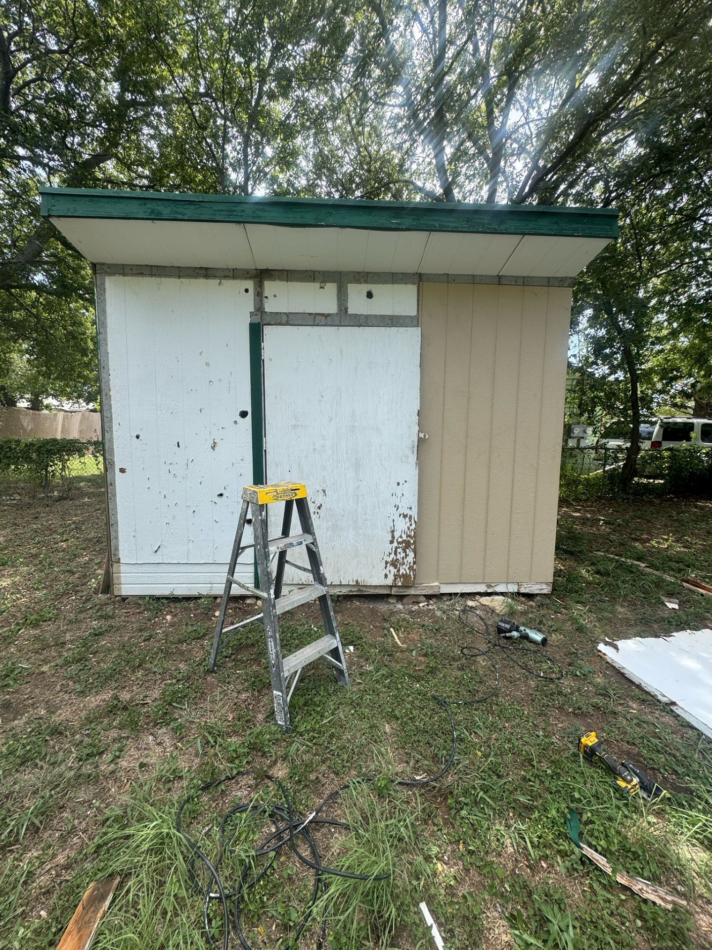 Small shed with white and tan doors, a ladder in front, and trees in the background.