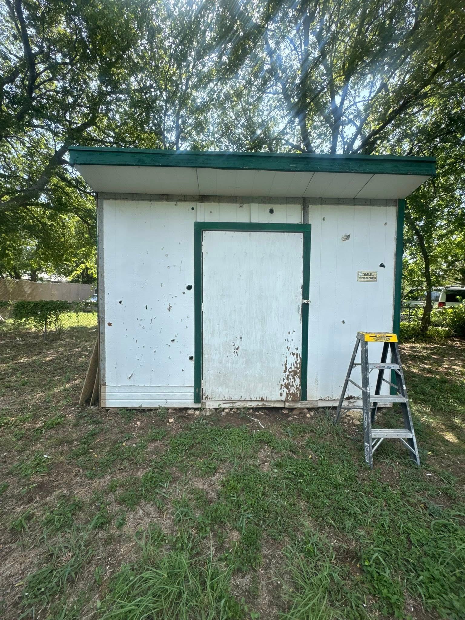Small white shed with green roof in a yard, with a step ladder beside the door.