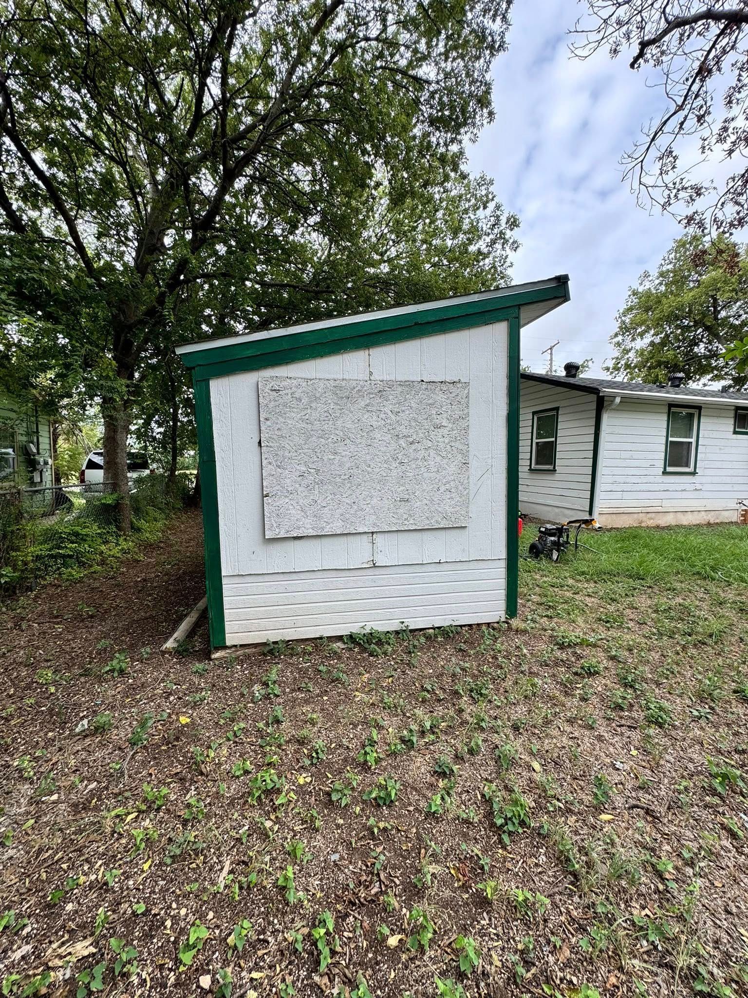 Small white shed with green trim and boarded window in a leaf-covered yard, beside a house and trees.