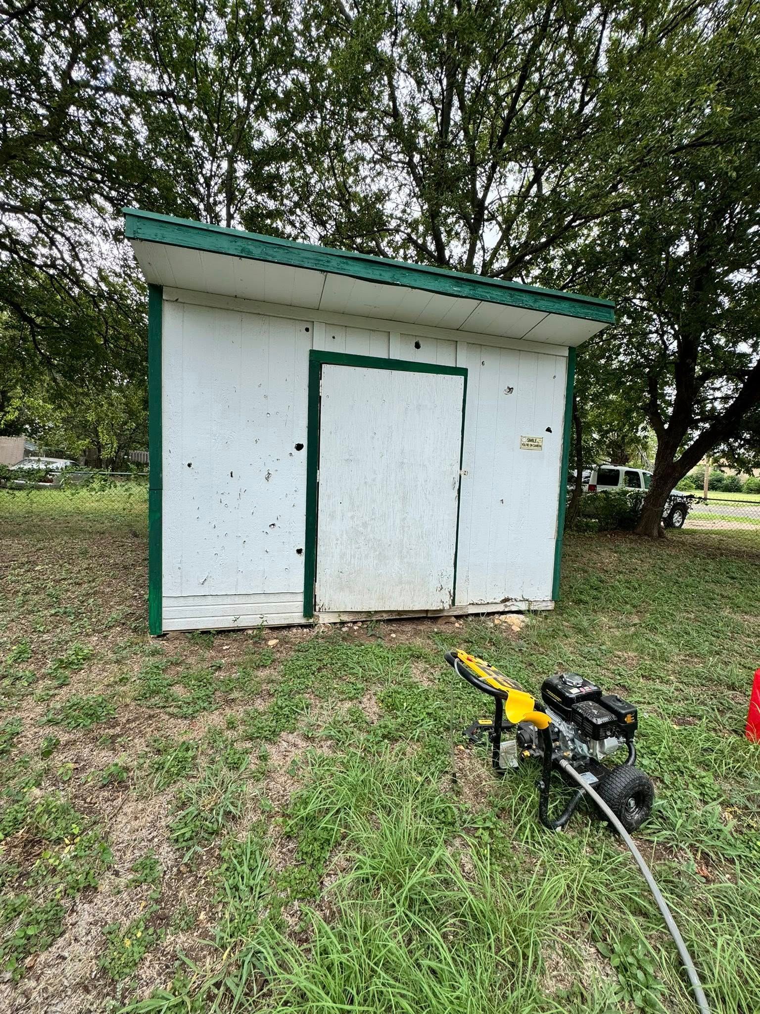 Small white utility shed in a grassy yard, with trees behind and a yellow tool on the ground.