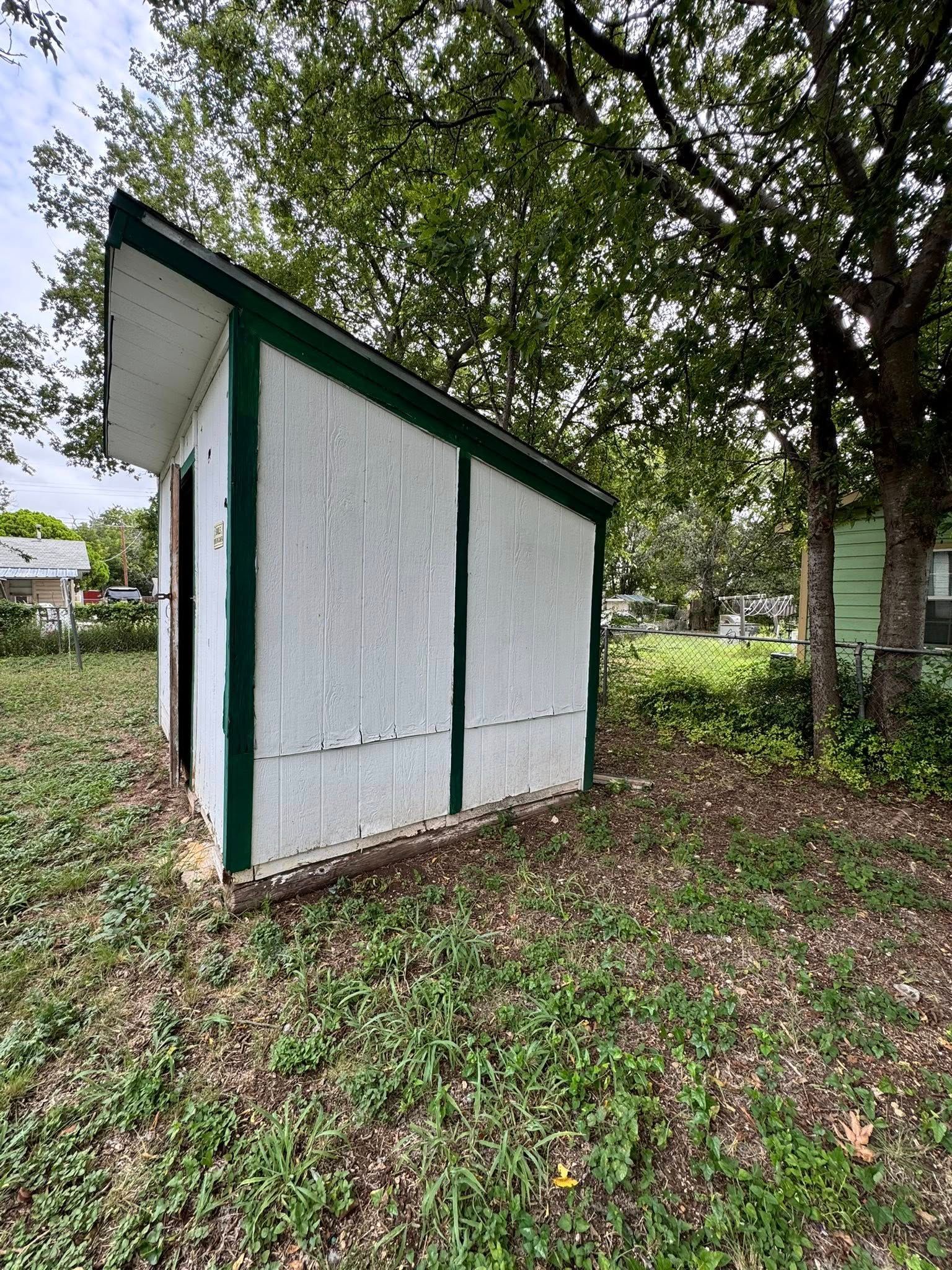 Small white shed with green trim on a grassy, leaf-covered lot under trees