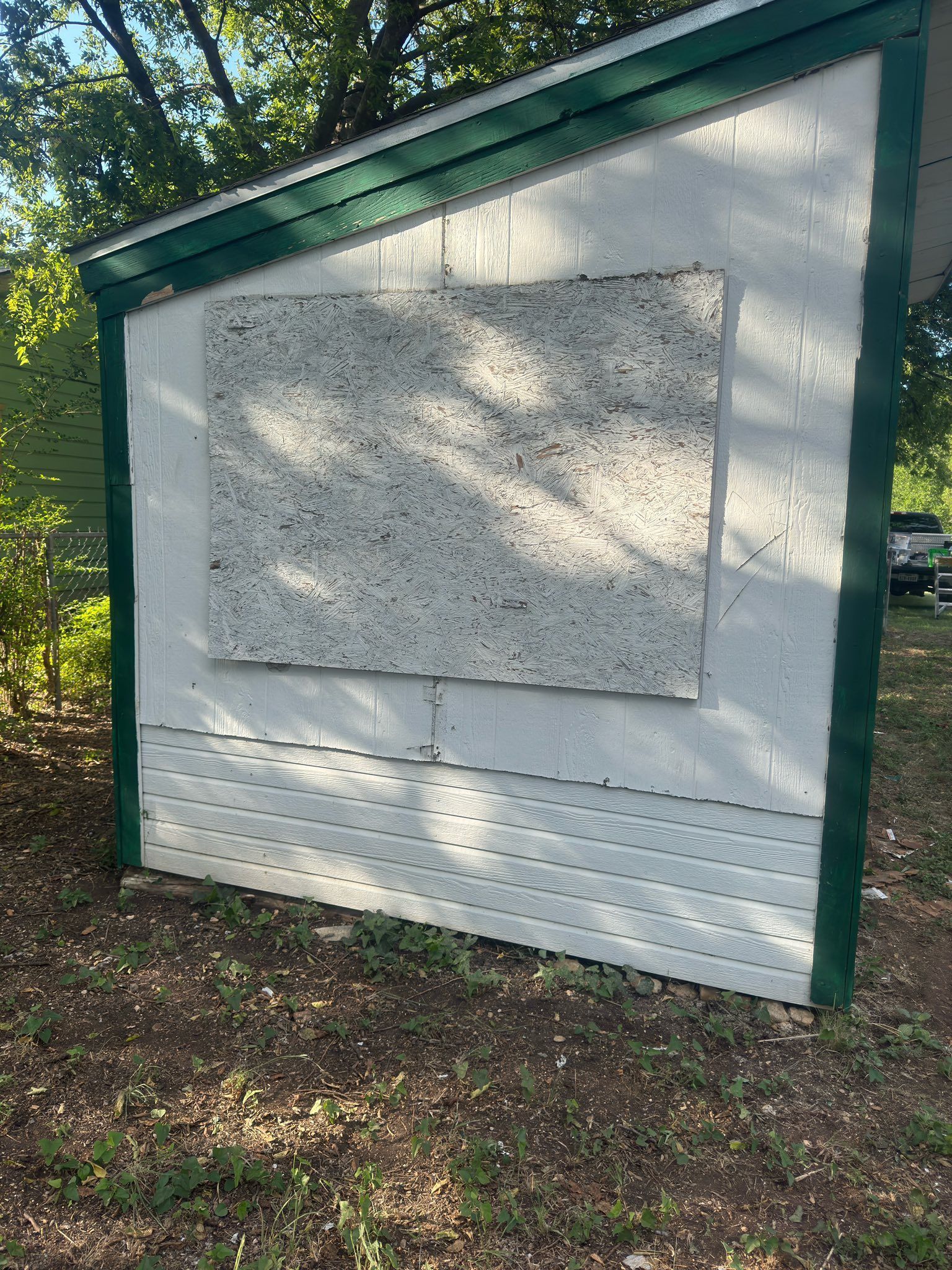 White shed with green trim and boarded window in a wooded outdoor area